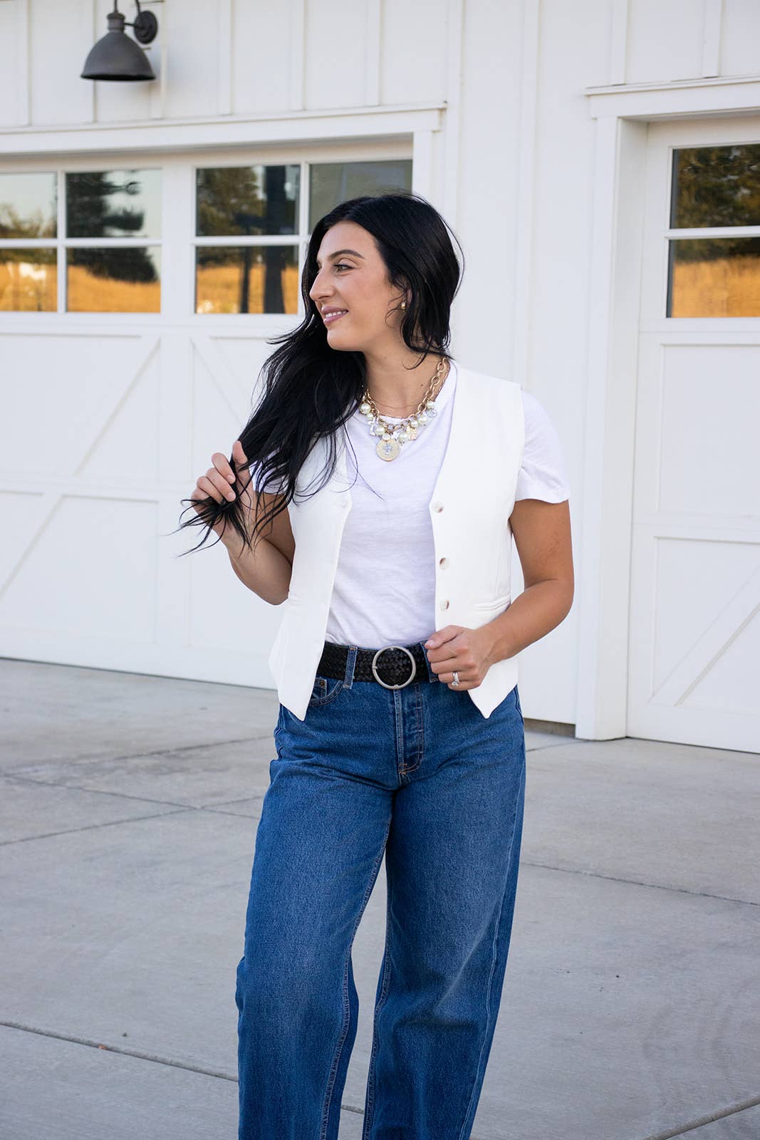 A woman with long dark hair, dressed in a white T-shirt, white vest, blue jeans, and a Ginger Blue Classic Wide Braided Leather Belt in Black, stands smiling outside in front of a white garage door.