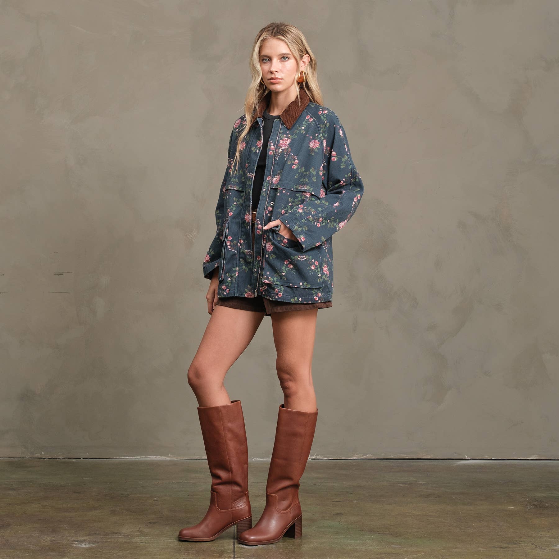 A woman with long blonde hair and a neutral expression stands indoors against a beige textured wall, wearing the RAISONNEL Floral Printed Utility Jacket (RJ1073), dark shorts, and knee-high brown boots.