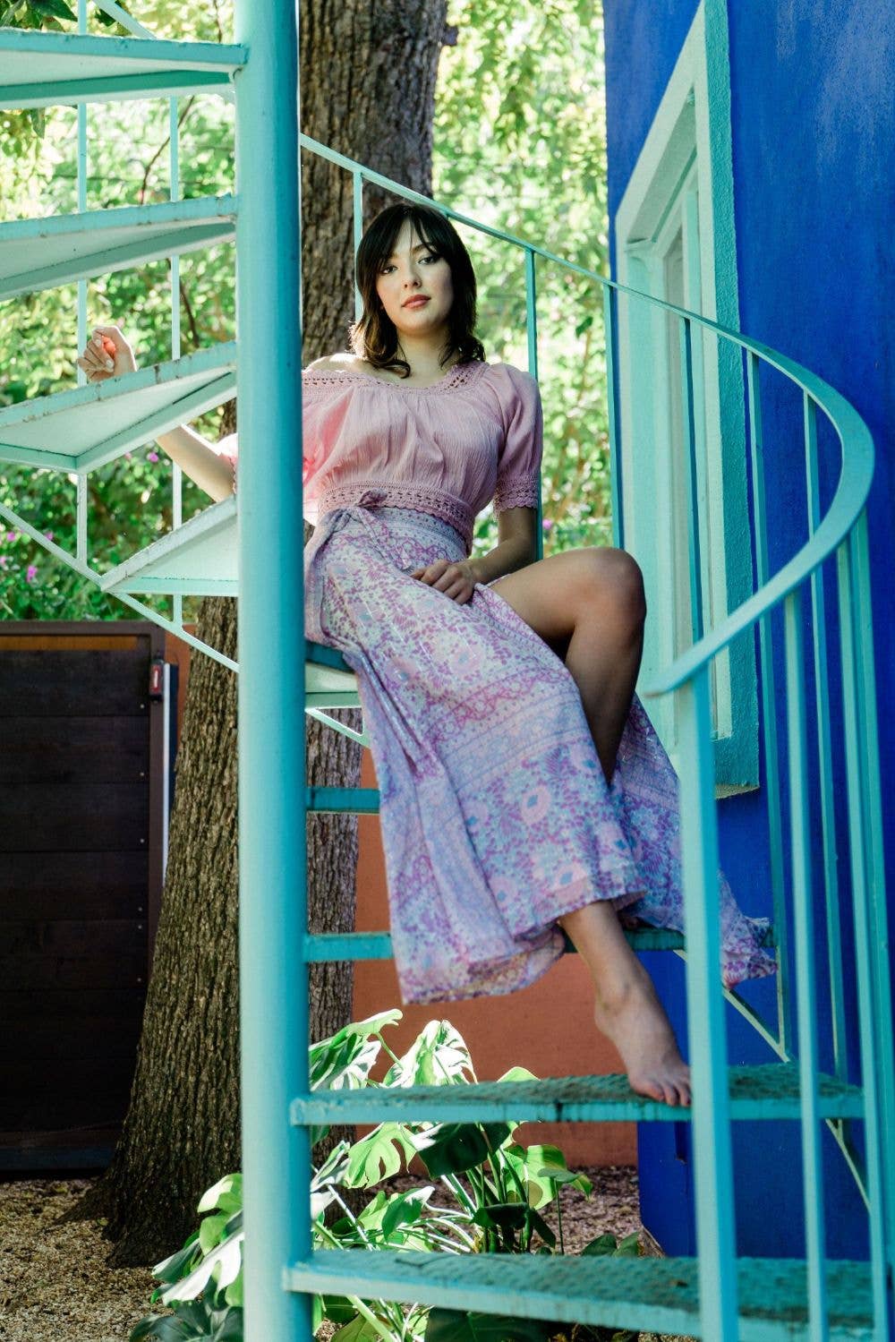 Woman sitting barefoot on turquoise spiral staircase wearing a patterned pink Glendora Wrap Skirt and blouse
