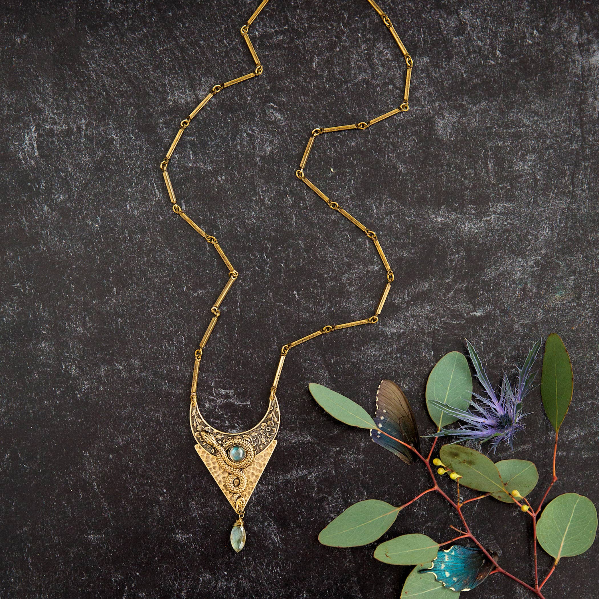 Moon of Ishtar Pendant Necklace with intricate brass design and blue gemstone, styled with green leaves on dark background