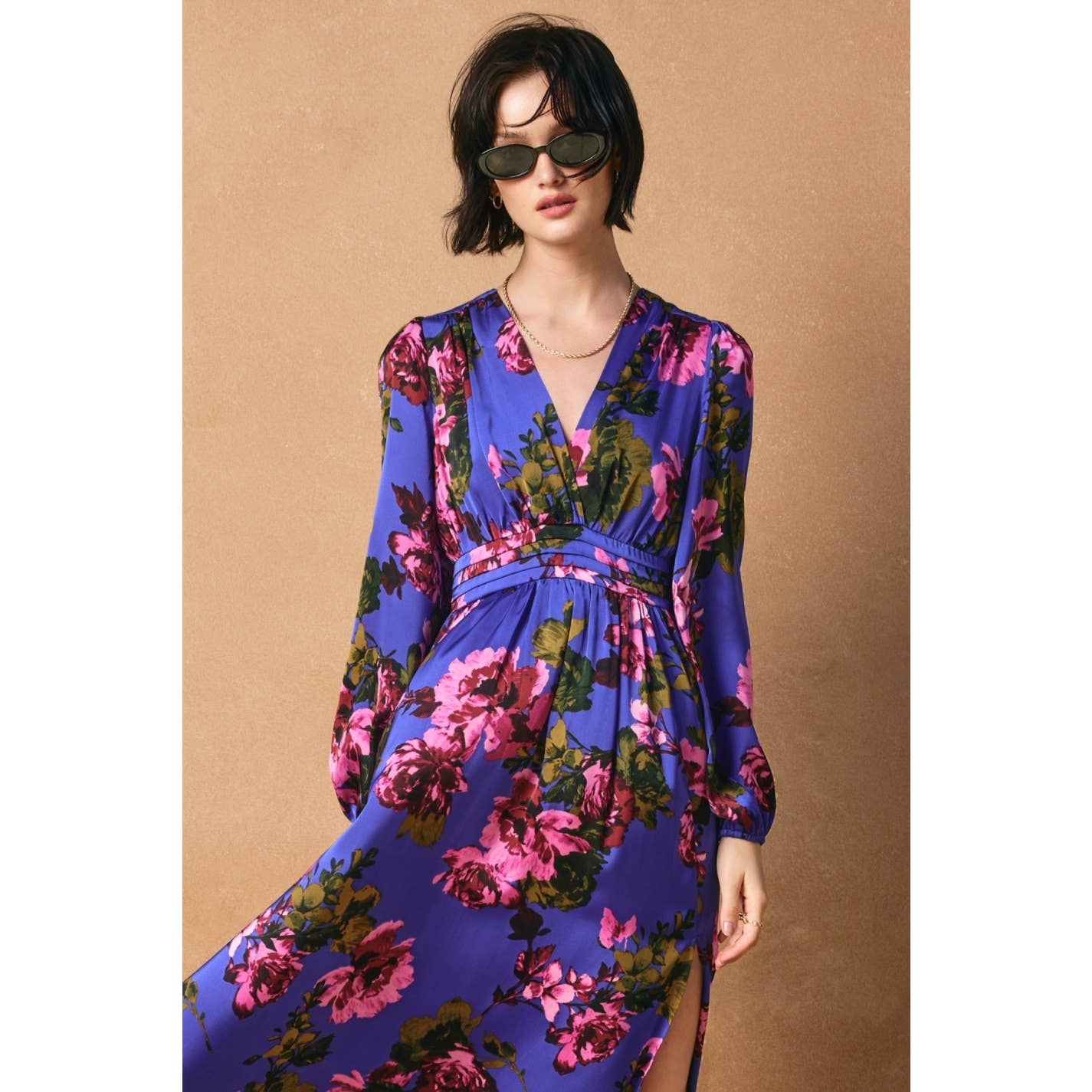 A woman in dark sunglasses and the Mesmerizing Essence Pleated Waist Maxi Dress by Dress Forum, featuring a blue floral print and pleated waist, stands against a tan background, her short tousled hair completing her confident look.