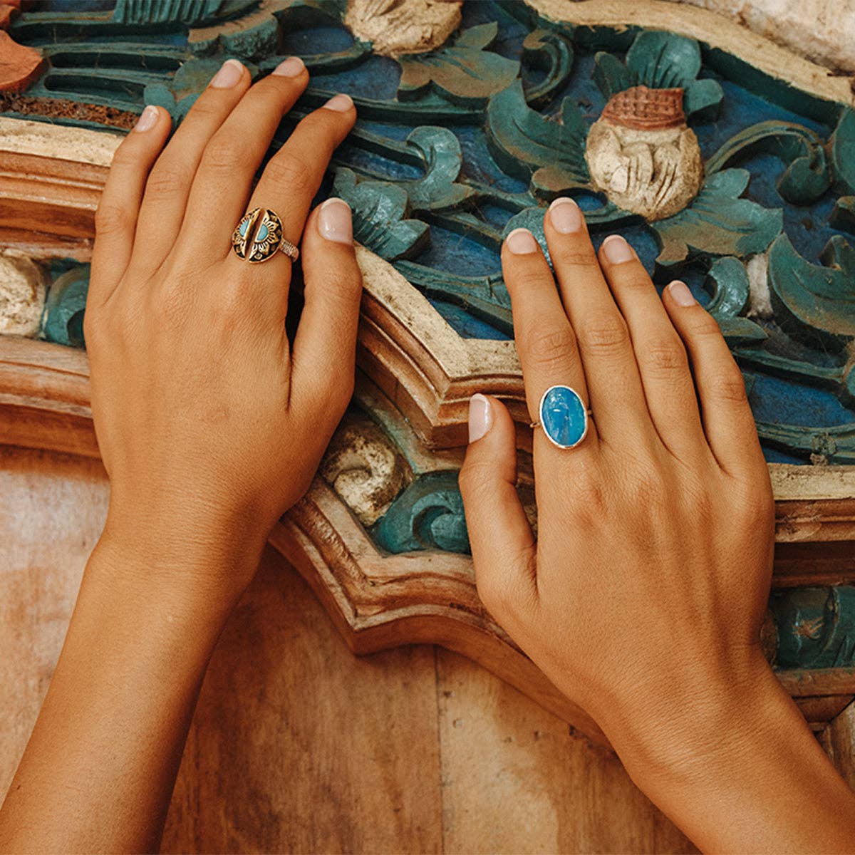 Two hands rest on an ornate painted surface. One wears a gold ring; the other showcases the bahgsu jewels Natural Mystic Ring - Turquoise, a bold, adjustable piece that catches the light with its striking turquoise stone.