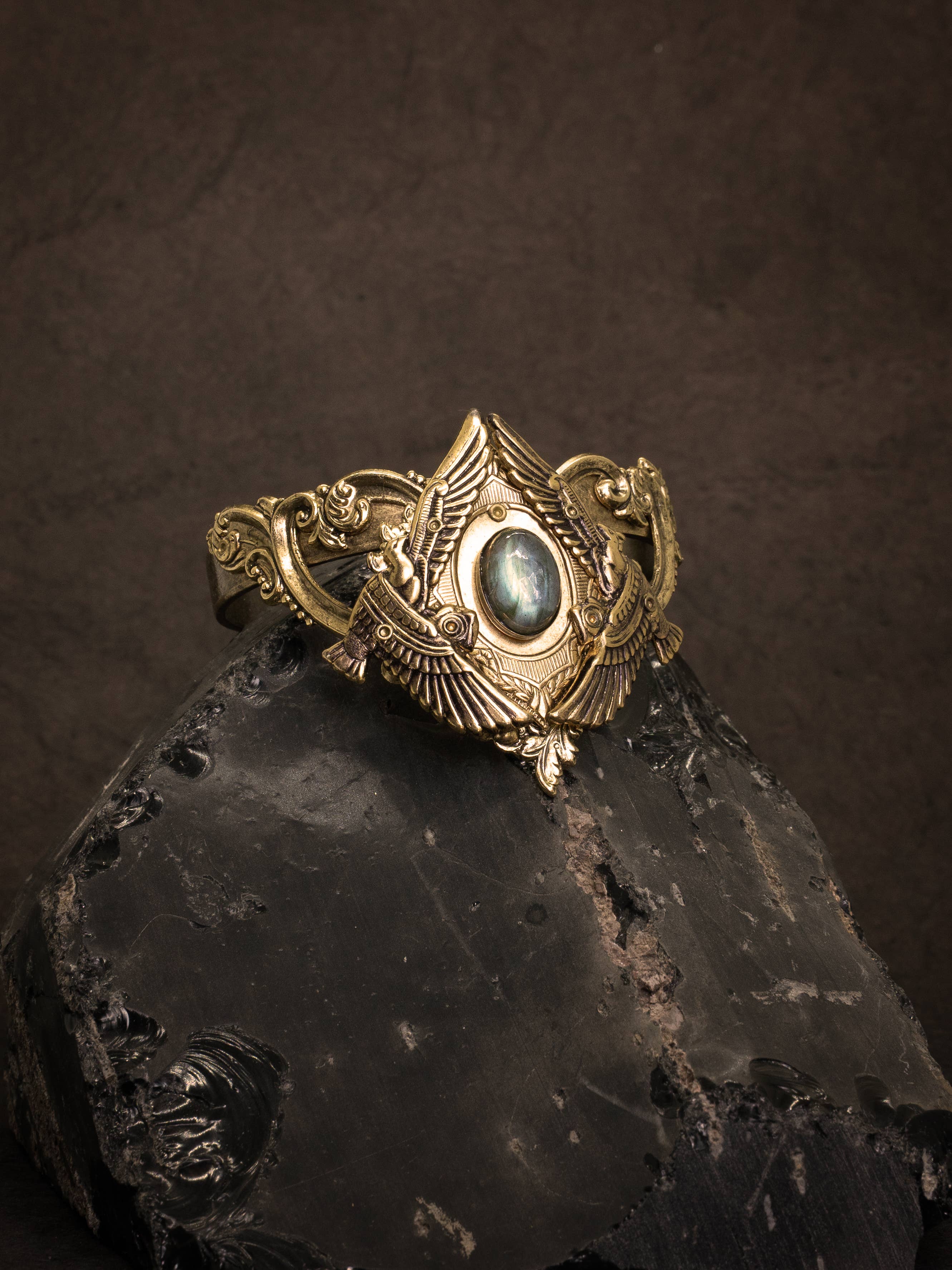 Nekhbet Shield Cuff featuring intricate winged design and central iridescent stone on dark textured background.