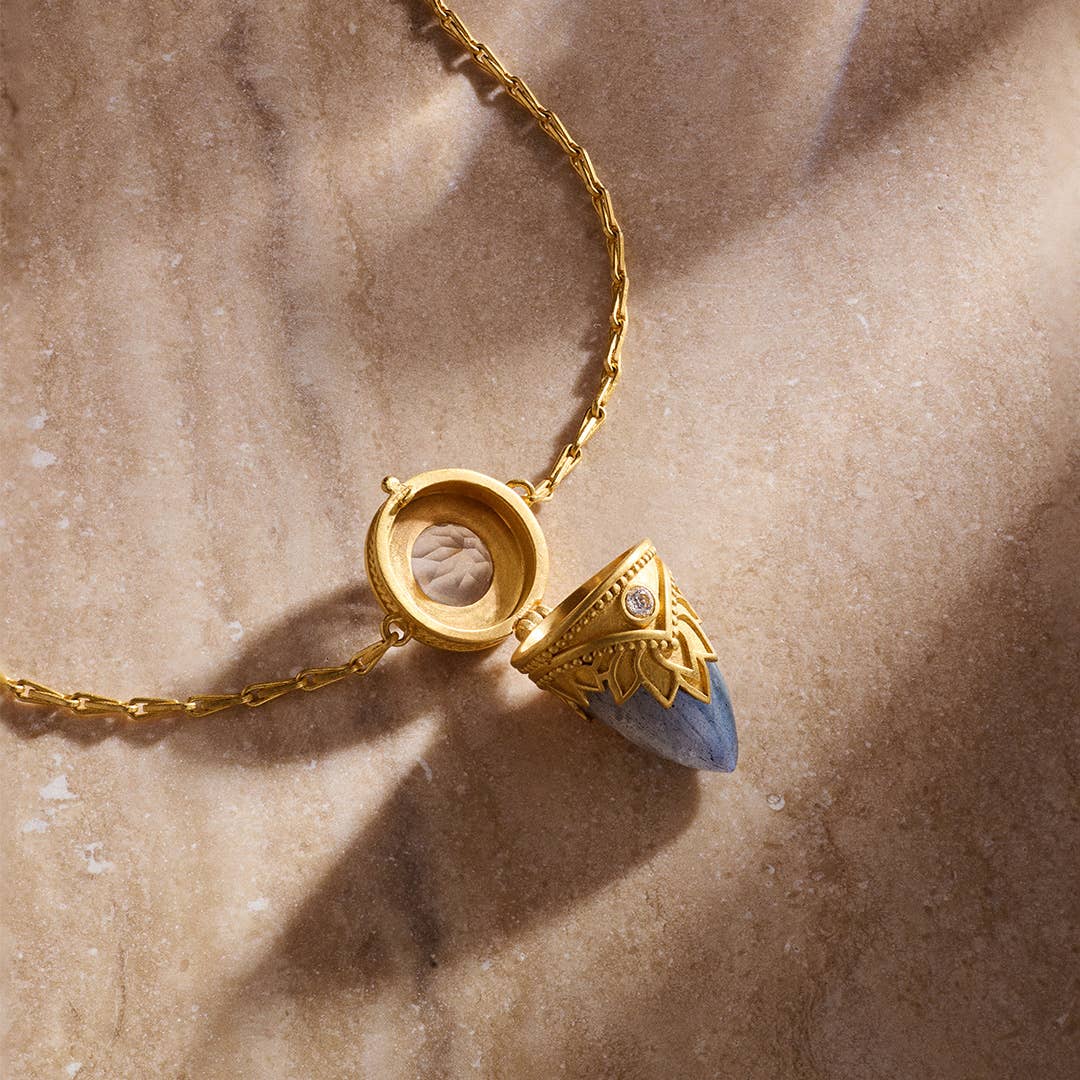The Satya Jewelry Guiding Spirit Labradorite Feather Locket Necklace - 18" rests on a beige marble surface, its gold and blue pendant partially open to reveal a hidden compartment, while soft light casts gentle shadows.