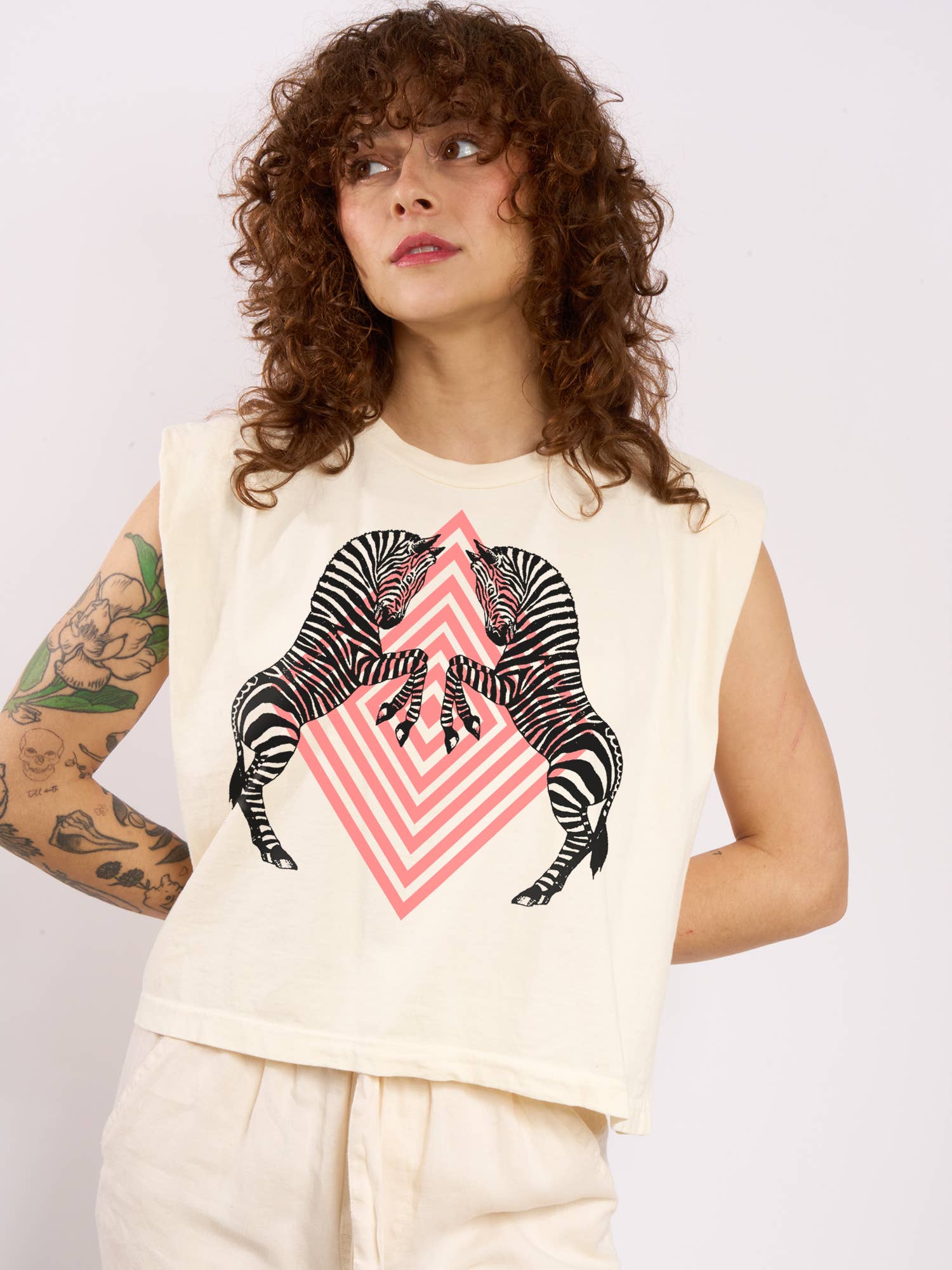 A person with curly hair and a tattooed arm wears the Supermaggie @ Faire Zebras Maisie Heavyweight Muscle Tee, a hand-printed cream cotton sleeveless top featuring two zebras facing each other over a pink diamond pattern.