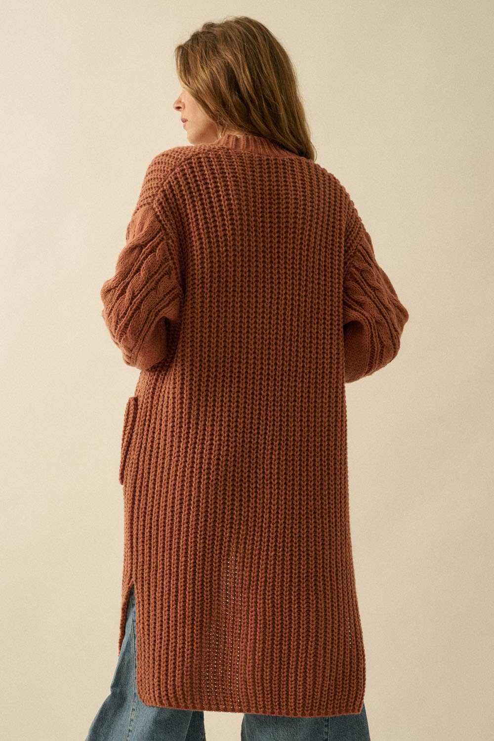 A woman with long brown hair, seen from behind, wears the Promesa USA Solid Rib Knit Open Front Cable Knit Cardigan in rust with blue jeans against a beige background—a cozy layering piece for any wardrobe.