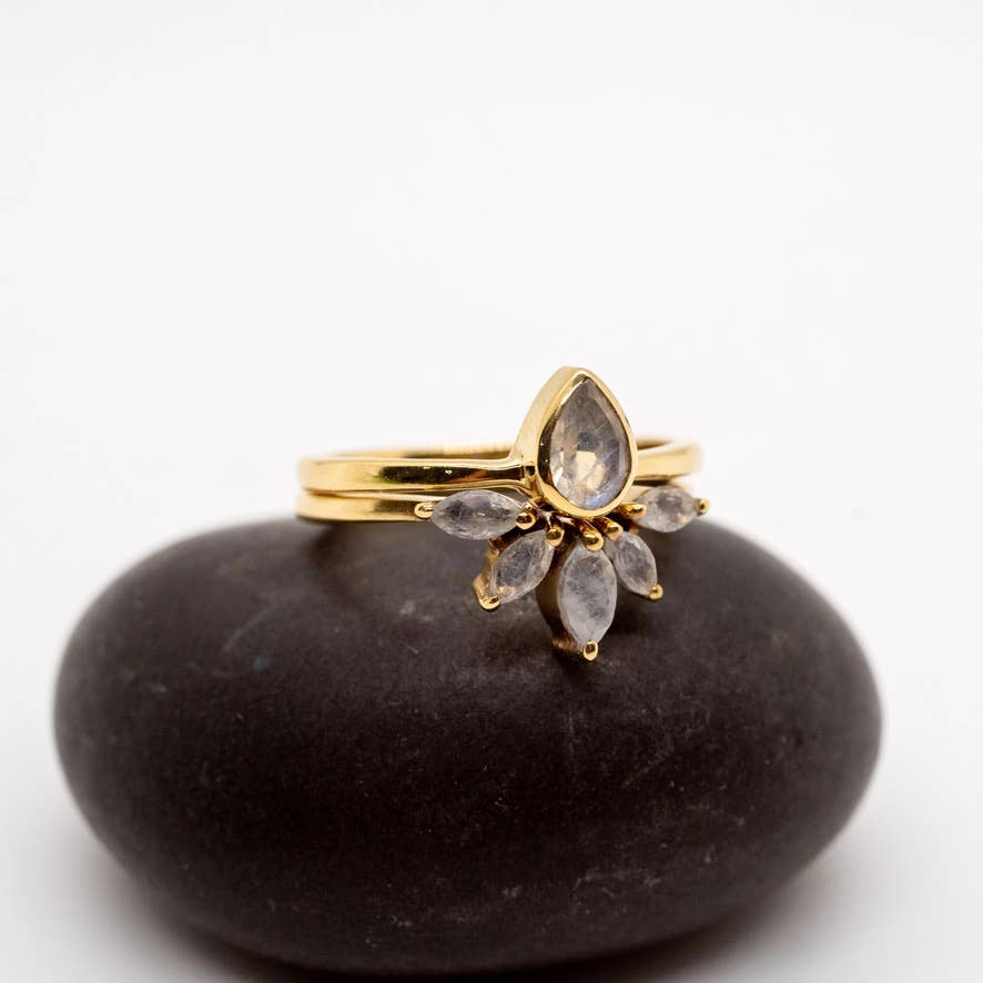 The Baizaar Brass Nesting Moonstone Ring Set features artisan craftsmanship with a teardrop-shaped moonstone and six marquise-cut stones, displayed on a smooth black oval rock against a white background.