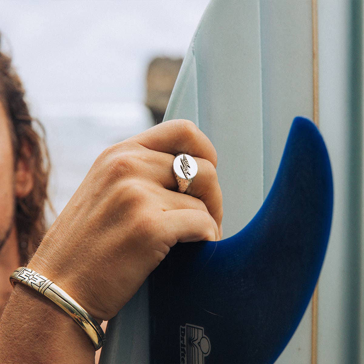Close-up of a hand wearing Medicine Wheel Cuff || Men and holding a surfboard fin by the beach.