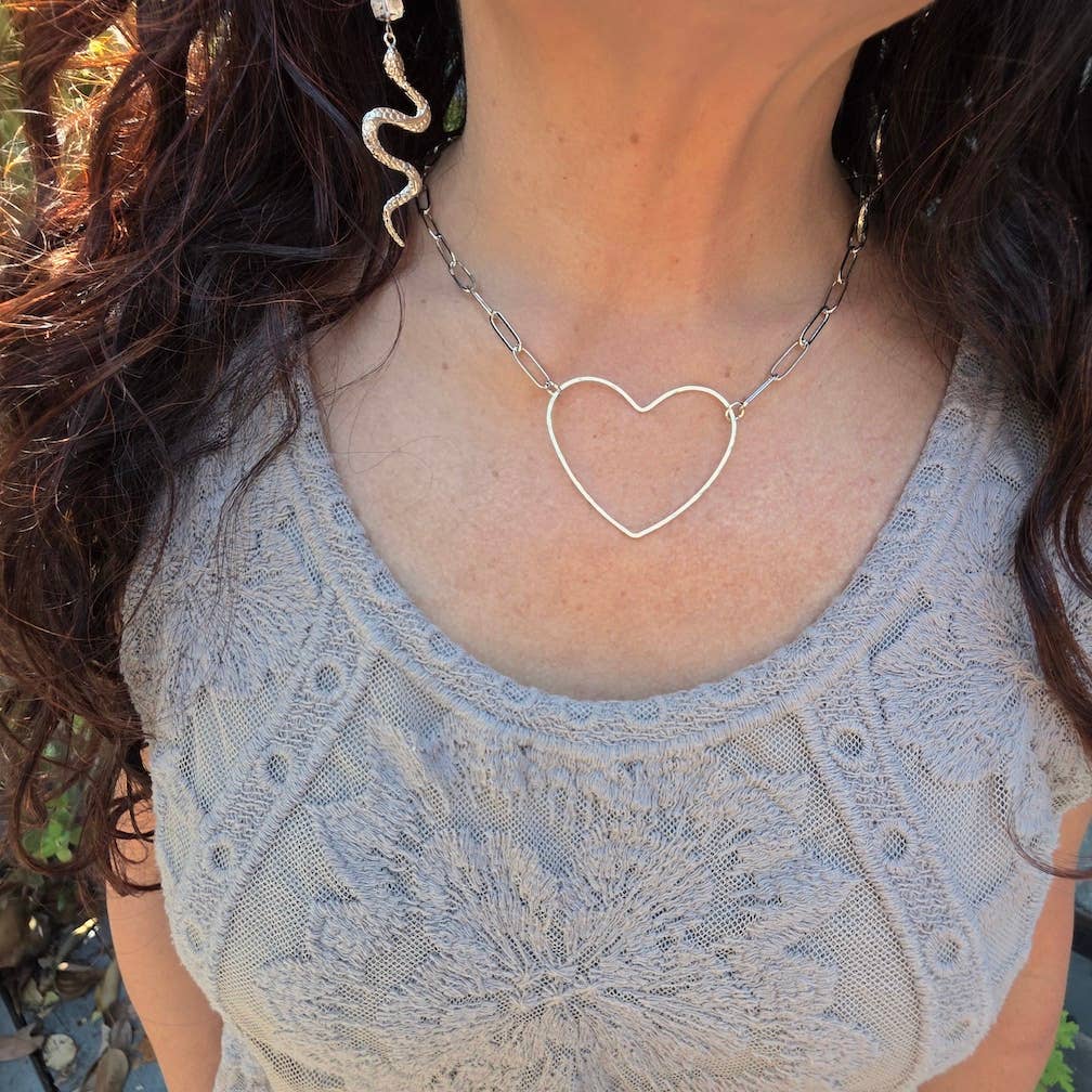 A woman shown from the shoulders up wears a light gray textured top, a Dreamers of Dreams Big Silver Heart - Paperclip Chain Necklace, and a silver snake-shaped earring.