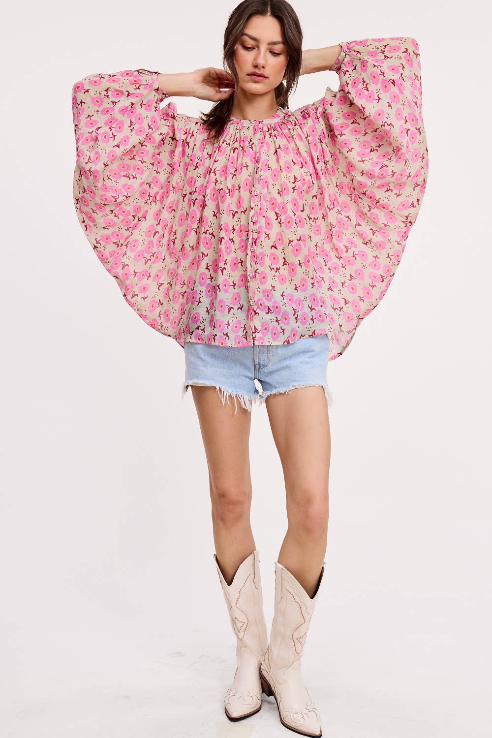 A woman models Ultramarine’s Meadow Breeze Linen Blouse, featuring loose pink floral prints and wide sleeves, styled with light blue denim shorts and white cowboy boots against a plain backdrop.