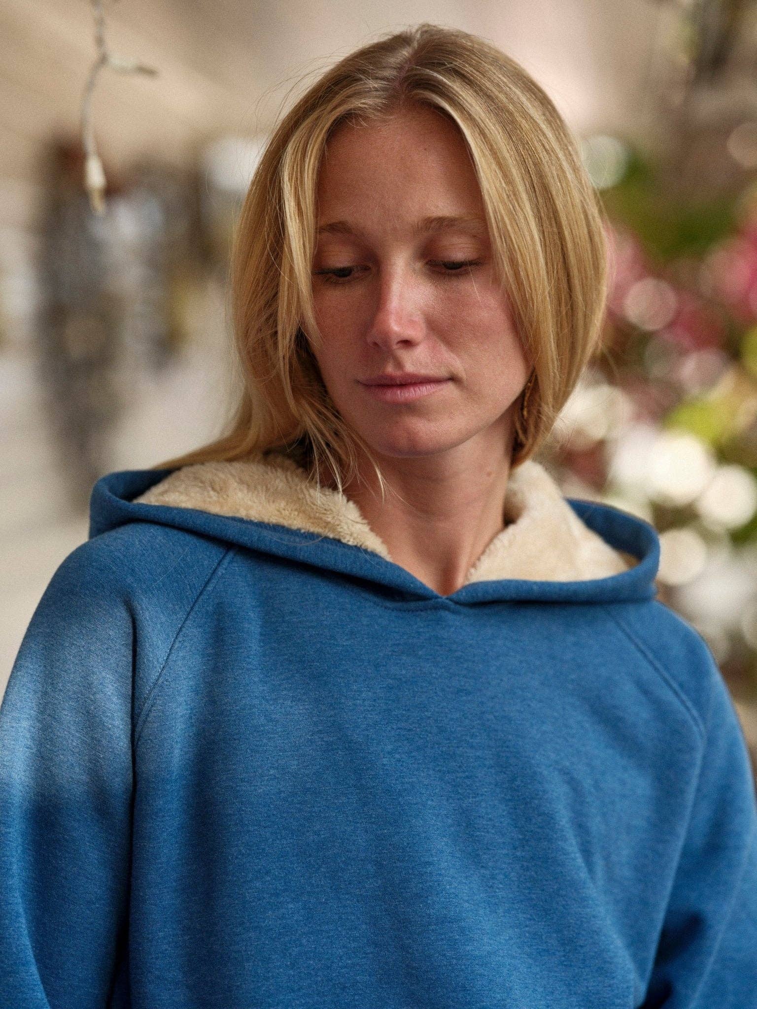 A person with blonde hair wears the Mollusk Cozy Sherpa-Lined Sweatshirt, featuring a blue exterior and light, fuzzy lining, while looking down with a neutral expression against a softly blurred backdrop of greenery and flowers.