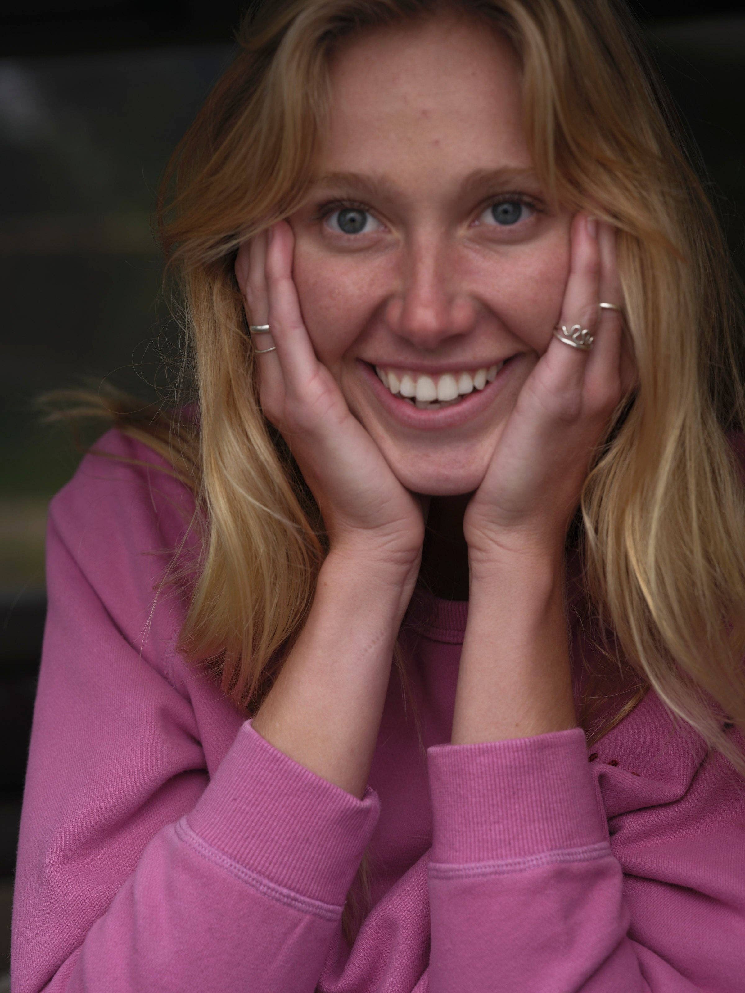 A young blonde woman with blue eyes smiles joyfully at the camera, resting her face in her hands while wearing the Mollusk Visions Crew sweatshirt and several rings.