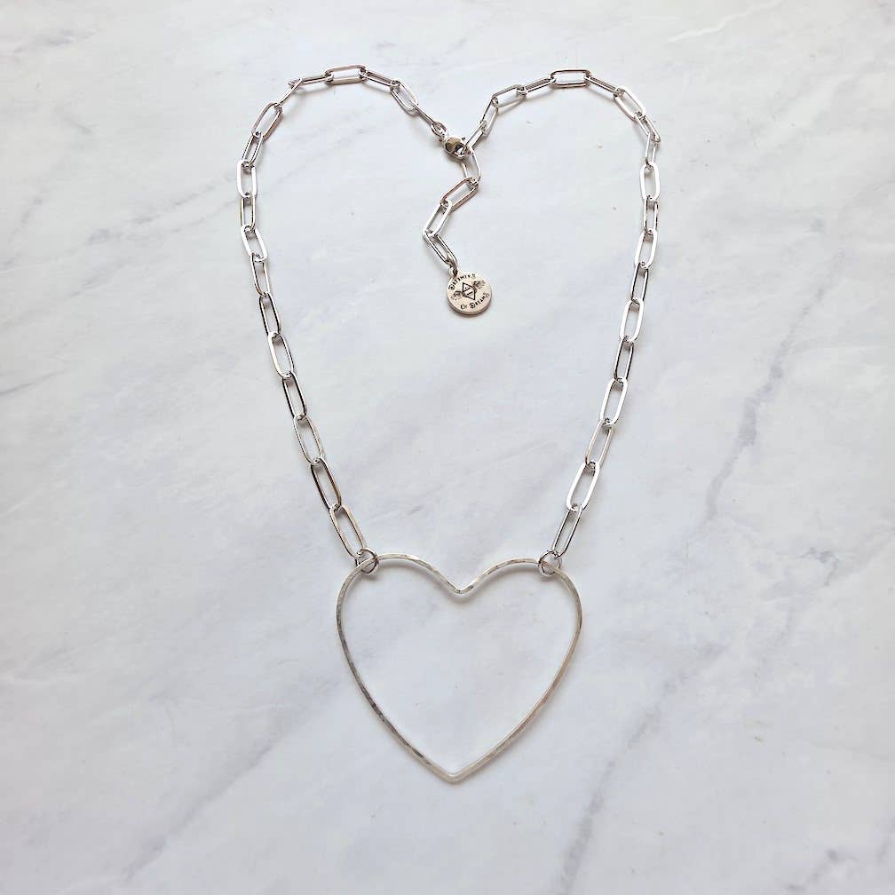 The Big Silver Heart -Paperclip Chain Necklace by Dreamers of Dreams features a silver-plated brass paperclip chain and a large hammered heart charm, with an extra small round charm near the clasp, displayed on light marble.
