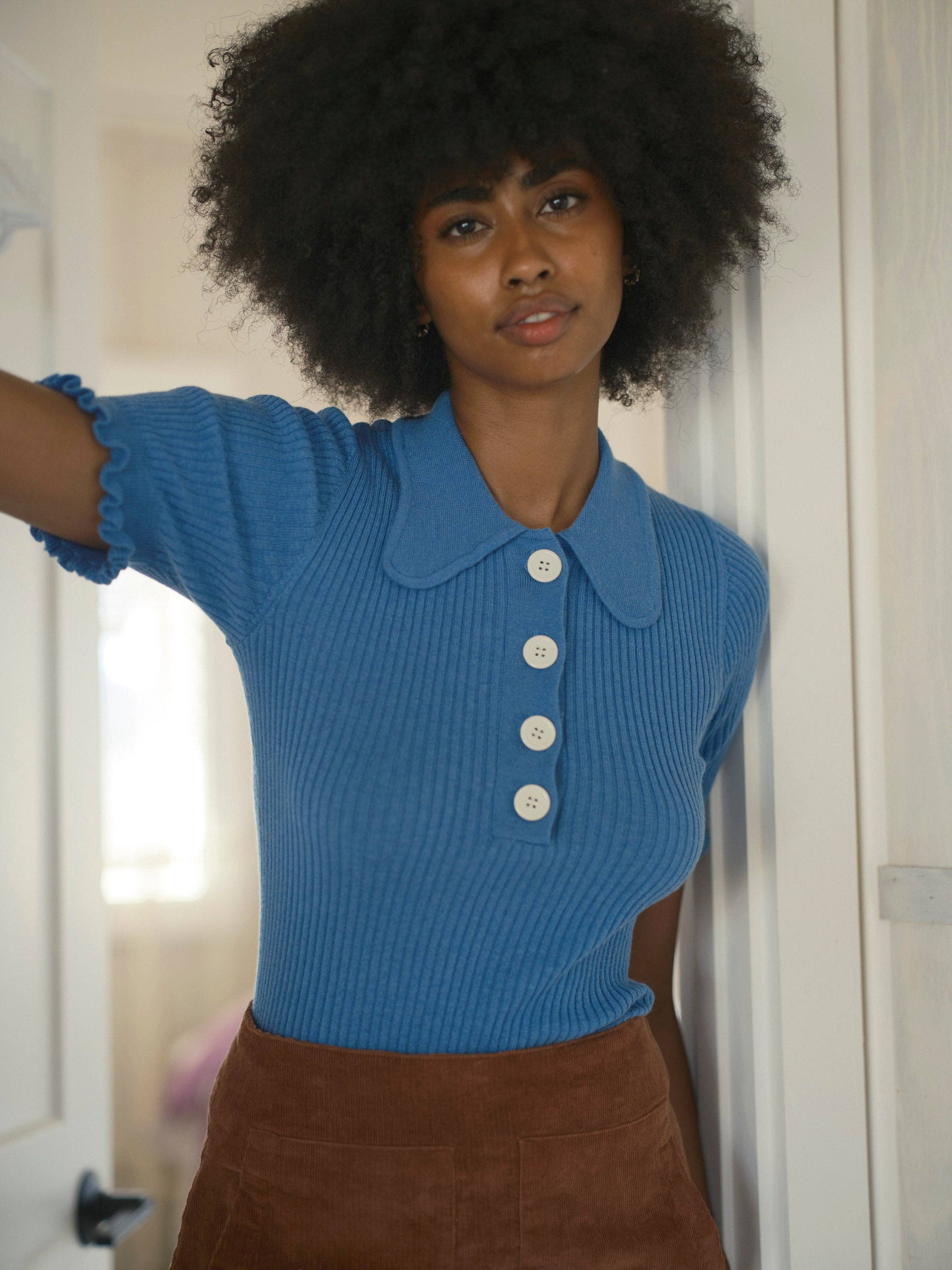 A woman with curly hair stands indoors, wearing the Mollusk Belle Tee—a vintage-inspired blue short-sleeve ribbed top with large white buttons and a pointed collar—paired with a brown skirt, looking into the camera with a neutral expression.