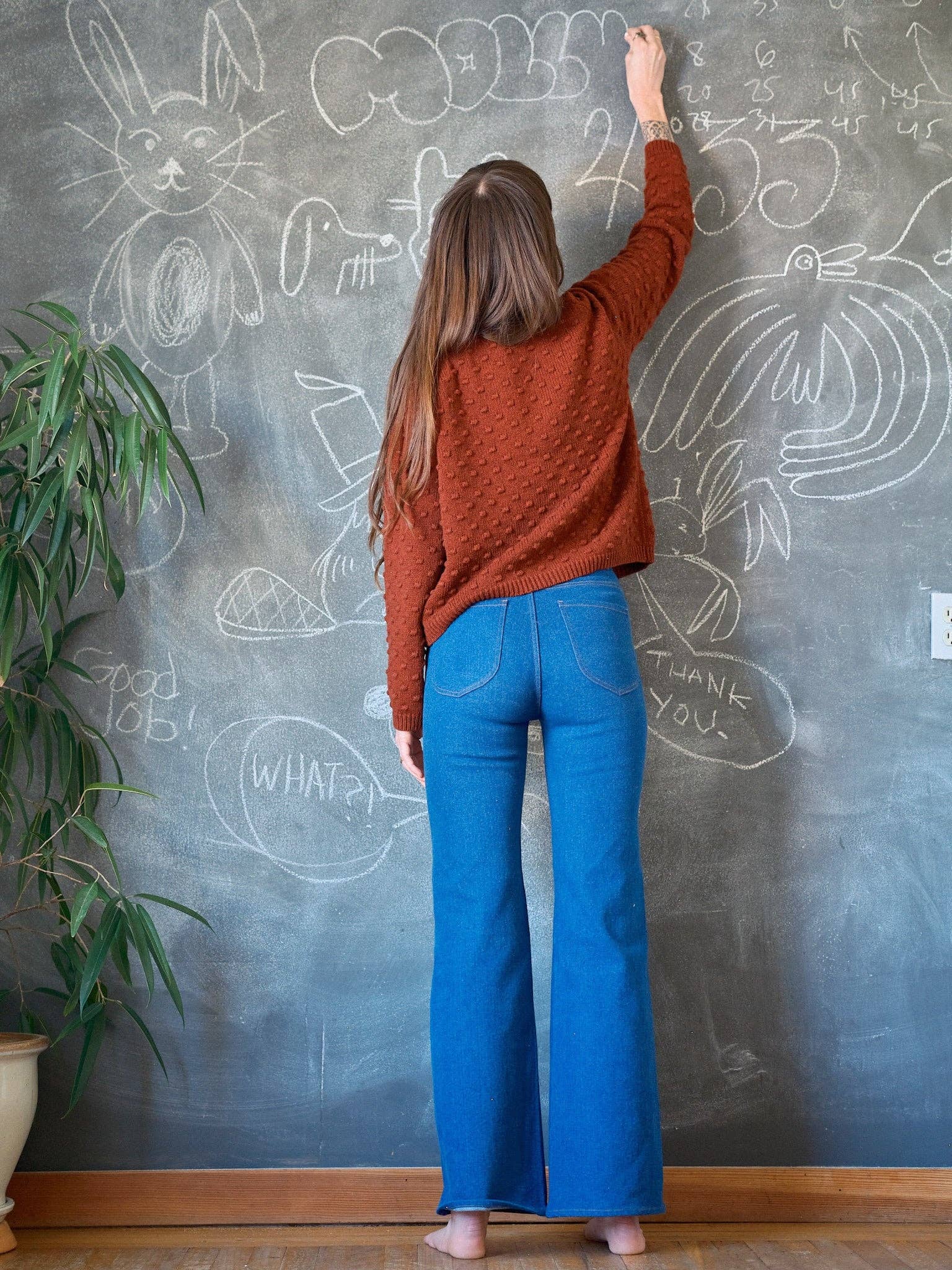 A woman with long brown hair, barefoot in blue jeans and a vintage-inspired Mollusk Pearl Sweater, draws animal doodles on a chalkboard wall. A green potted plant sits to her left.