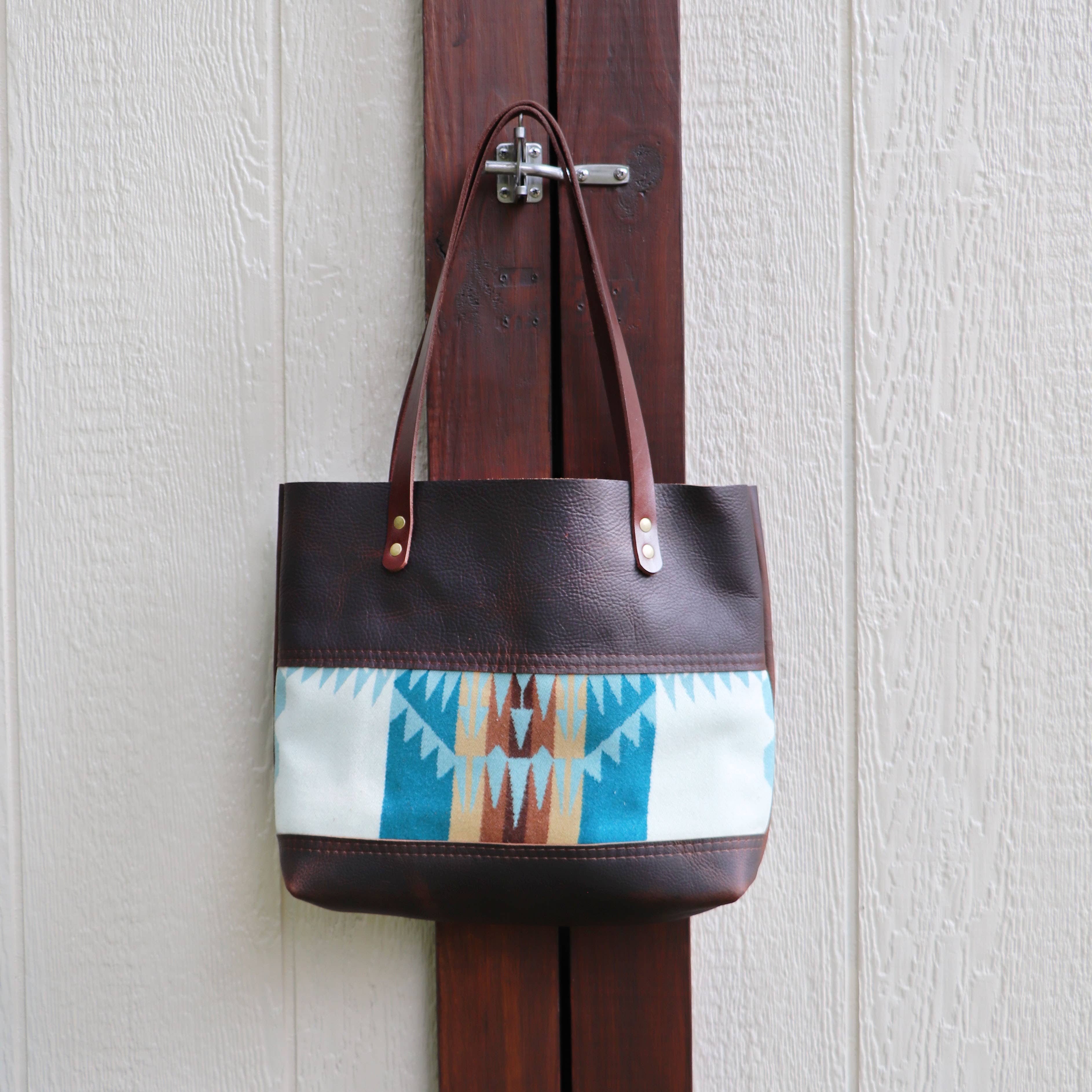 The Hawks & Doves Wool Tote Short is a brown oil tanned leather bag with long handles and a Southwestern blue, white, and brown fabric panel, shown hanging on a wooden door latch against a textured light wall.