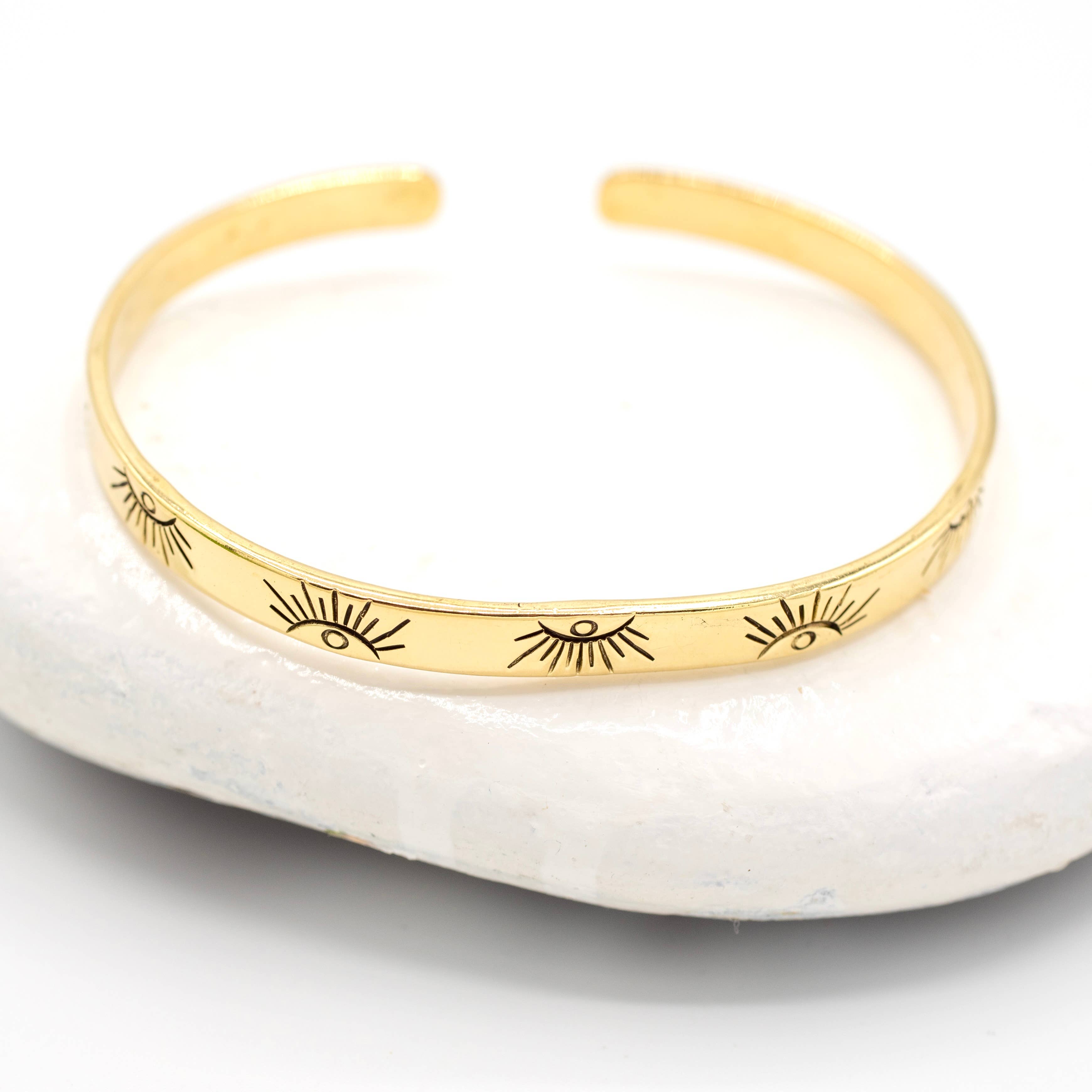 The Baizaar Brass Eye Bangle, a handmade bracelet with engraved eye and lash motifs, is shown resting on a white stone against a simple background.
