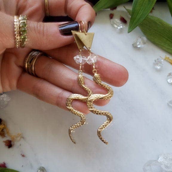 A hand holds Dreamers of Dreams Gold Year of the Snake Earrings, featuring crystal beads and triangle tops, surrounded by green leaves and clear crystals on a white surface.