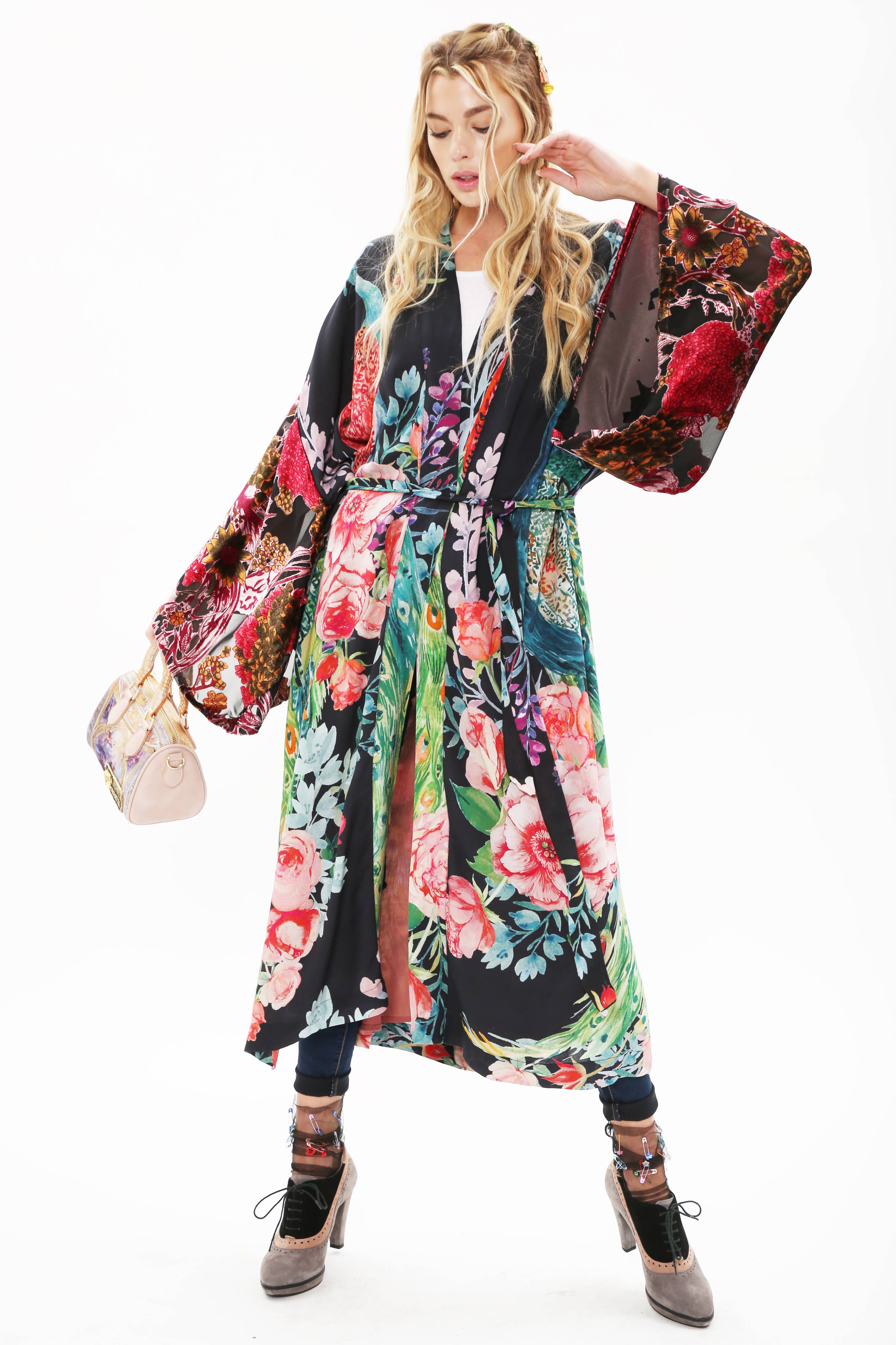 A woman models the Aratta Sweet Fantasy Kimono with wide burnout velvet sleeves, paired with blue jeans and high heels. She holds a small light pink handbag, her long wavy blonde hair styled in braids.