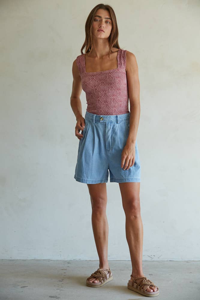 A woman stands indoors against a light wall, wearing By Together’s Knit Modal Nylon Spandex Seamless Jacquard Tank, high-waisted light blue shorts, and tan slip-on sandals. She has loose straight hair and a relaxed, neutral expression.