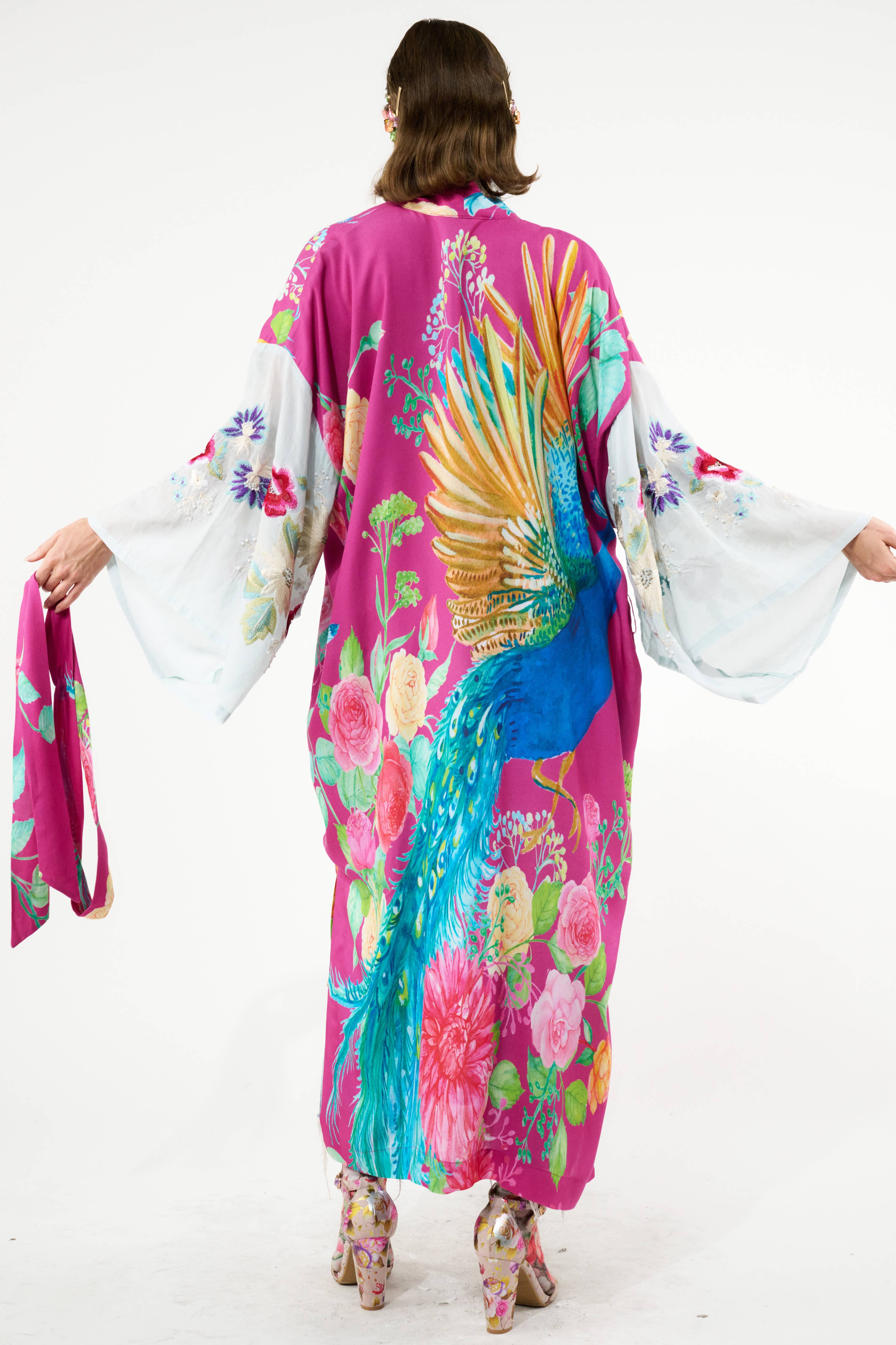A person stands facing away, wearing the Aratta Marie Antoinette Kimono with wide embroidered floral sleeves and a bold blue and gold peacock design on the back, styled with matching high-heeled shoes for a bohemian look.