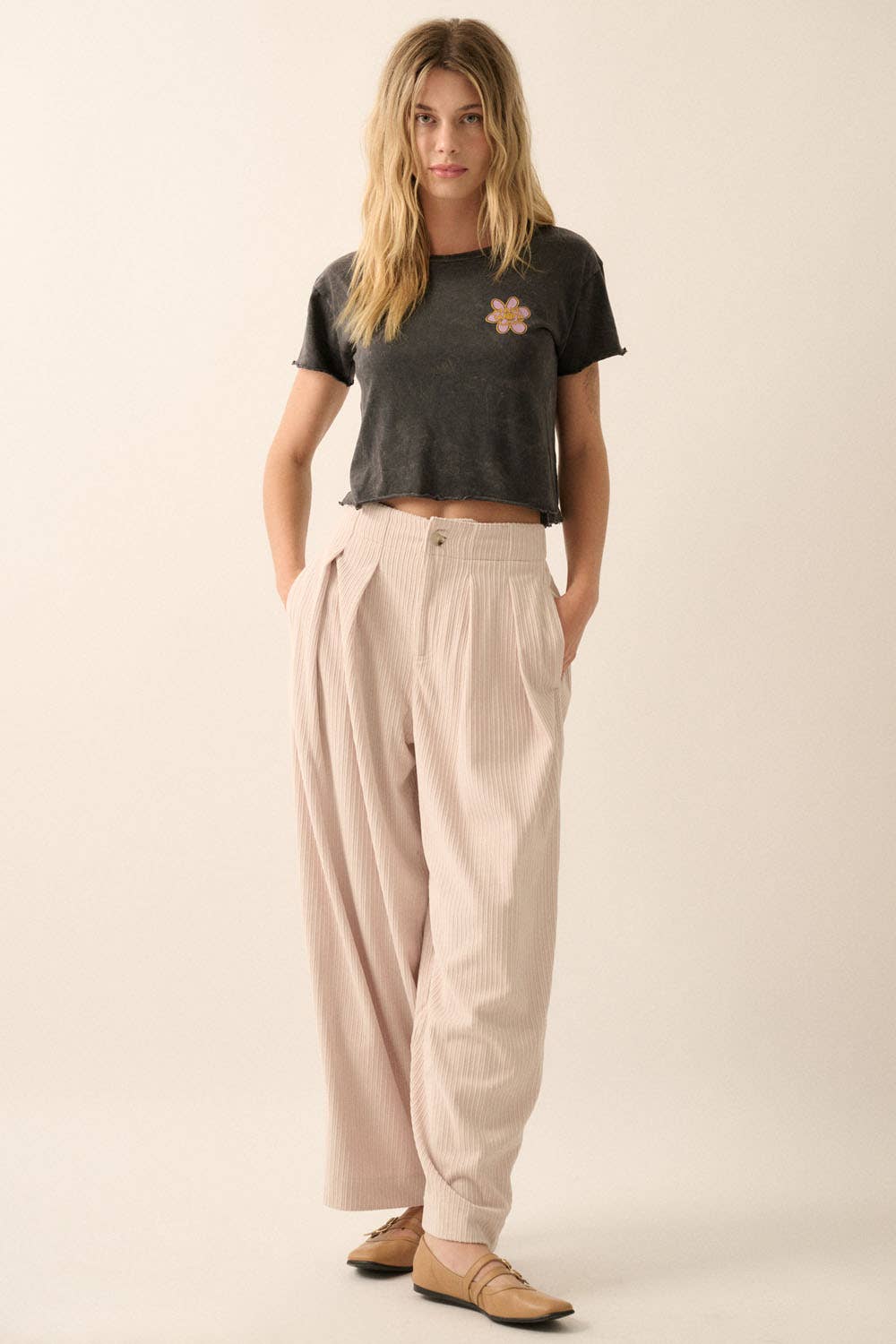 A woman stands with hands in pockets, wearing a black cropped t-shirt with a flower graphic, Promesa USA Velvet Corduroy Pleat-Front Wide-Leg Pants in beige, and tan pointed-toe flats against a plain background.