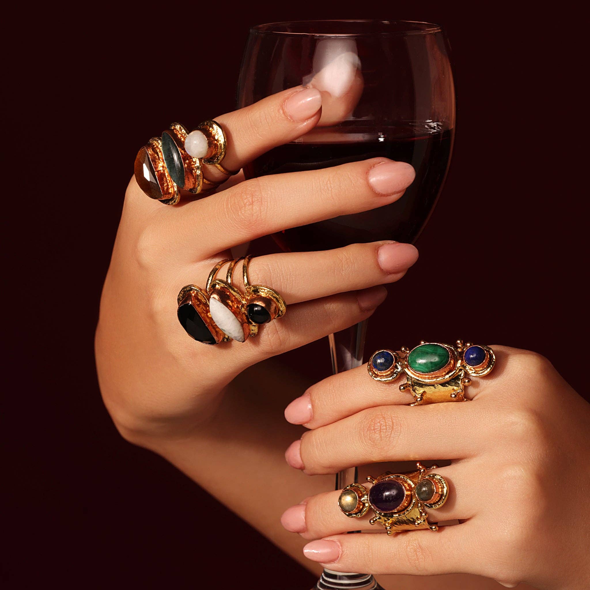 Hands adorned with multiple rings holding a glass of red wine featuring the Aurora Luxe Gemstone Ring