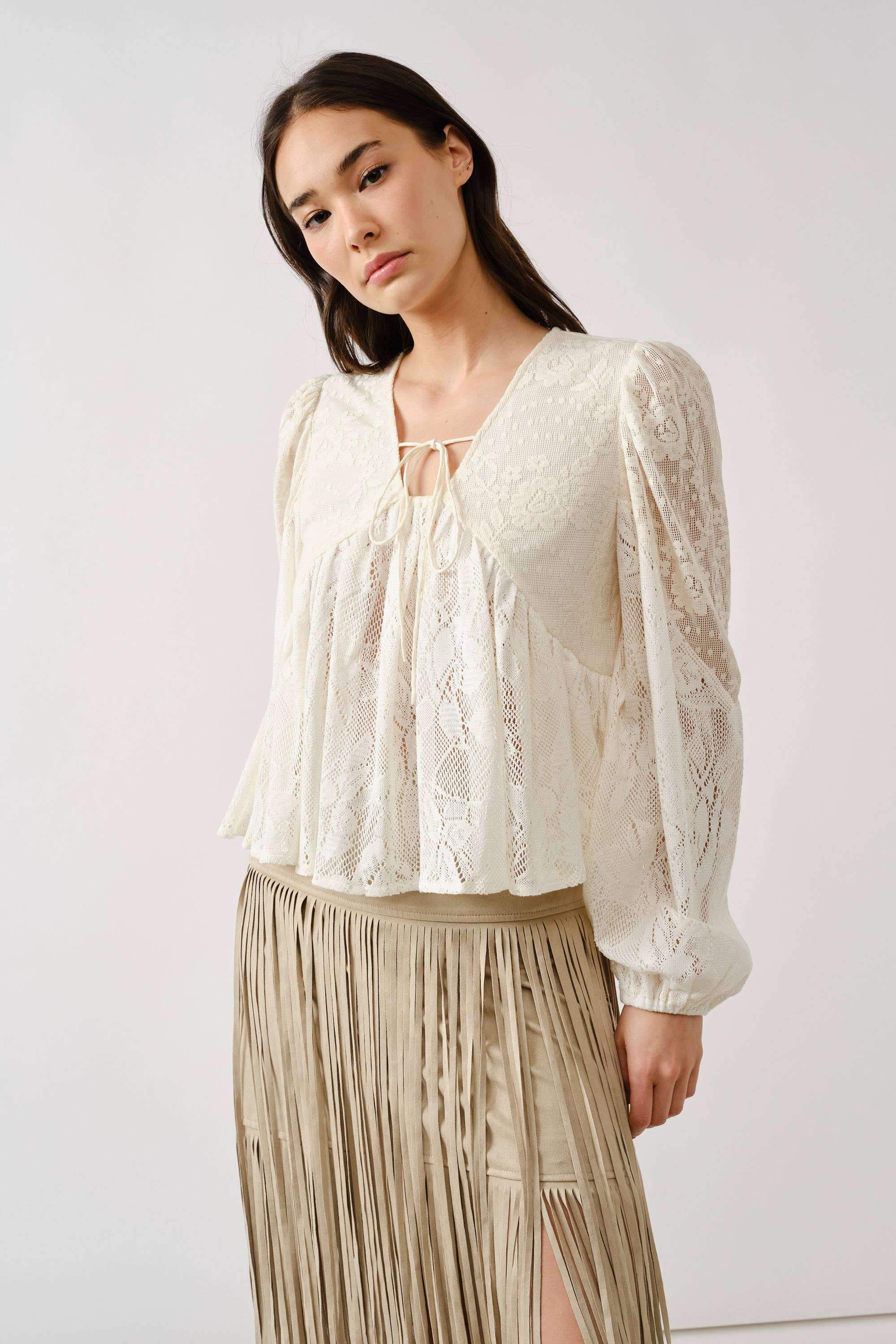 A woman wears the Aaron & Amber Long Sleeve Lace Babydoll, a cream floral lace blouse with a tie neckline, paired with a beige fringed skirt, standing against a plain light background.