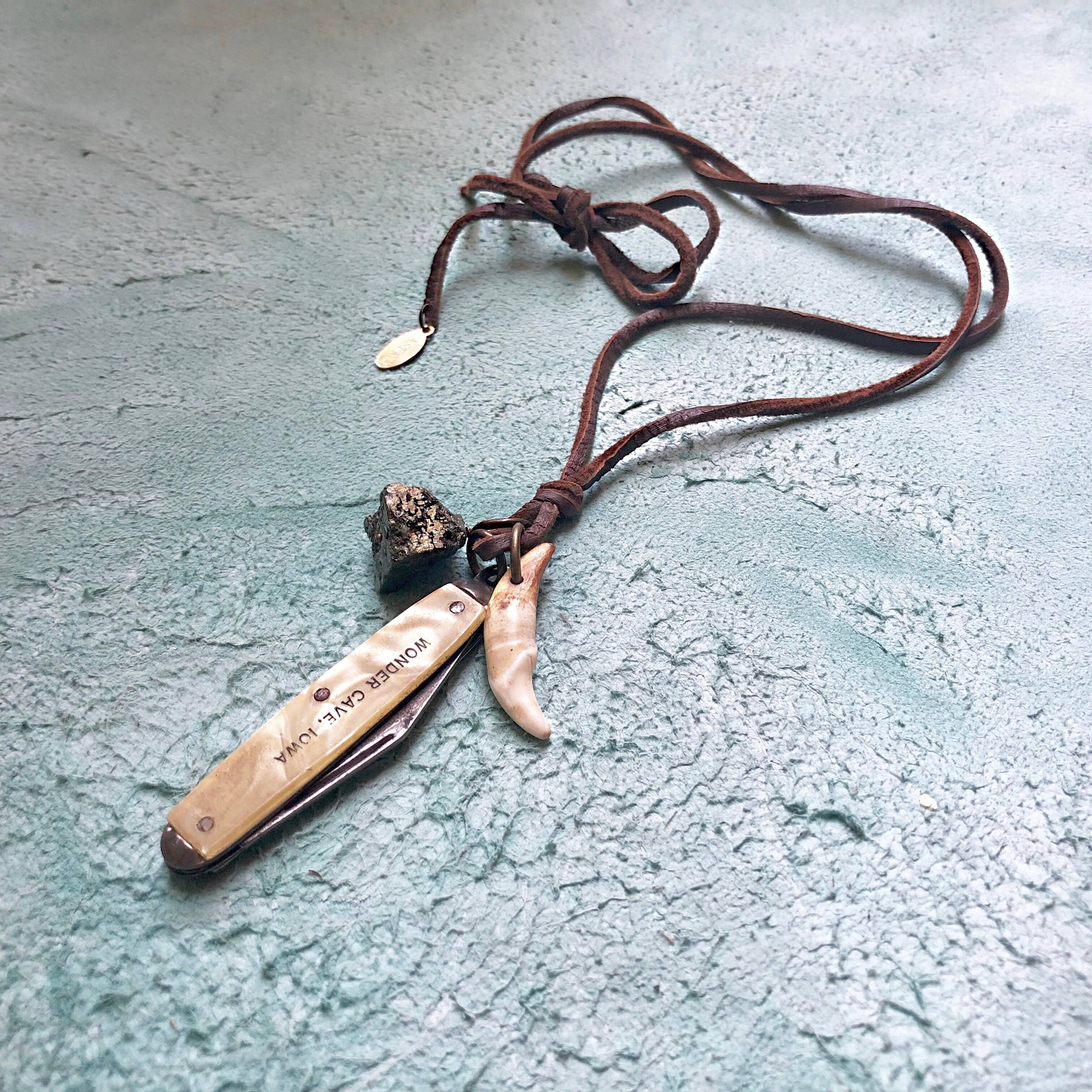 The ASTALI Vintage Pocket Knife & Coyote Tooth Necklace features a pyrite stone and deerskin leather cord, displayed on a textured light blue surface.