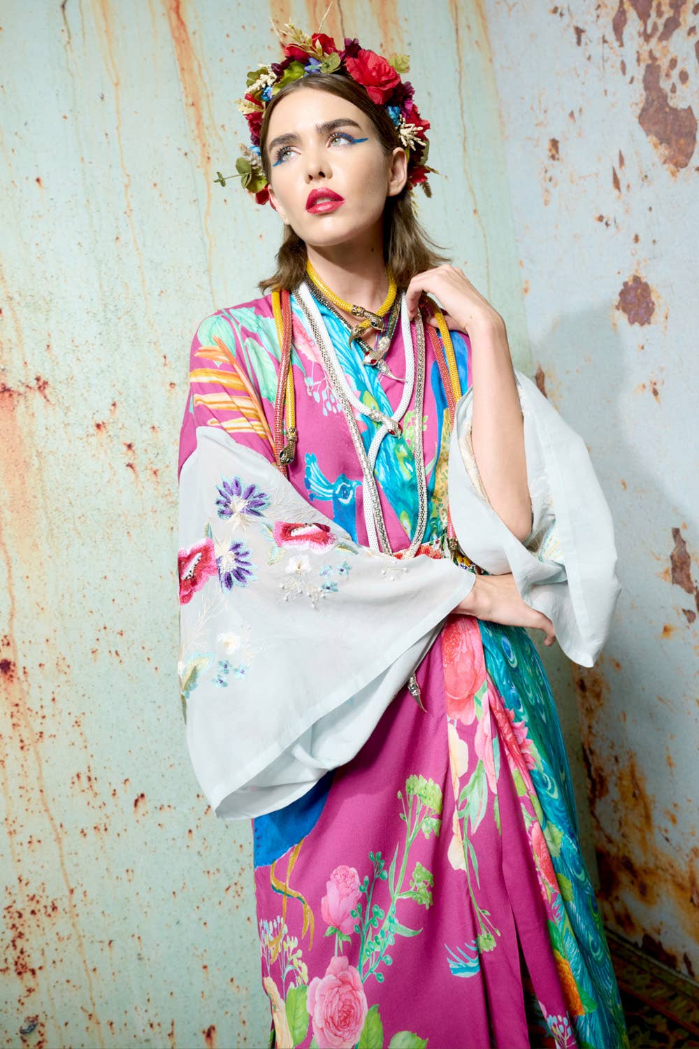 A woman in the Aratta Marie Antoinette Kimono, with embroidered floral sleeves and a flower crown, stands by a rusty wall. She gazes upward, red lipstick and colorful necklaces completing her unique bohemian look.