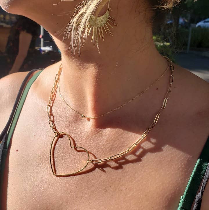 A person wears Dreamers of Dreams’ Big Gold Heart | Paperclip Chain Necklace layered with gold sunburst earrings and a green spaghetti strap top, sunlight casting delicate shadows on their skin.