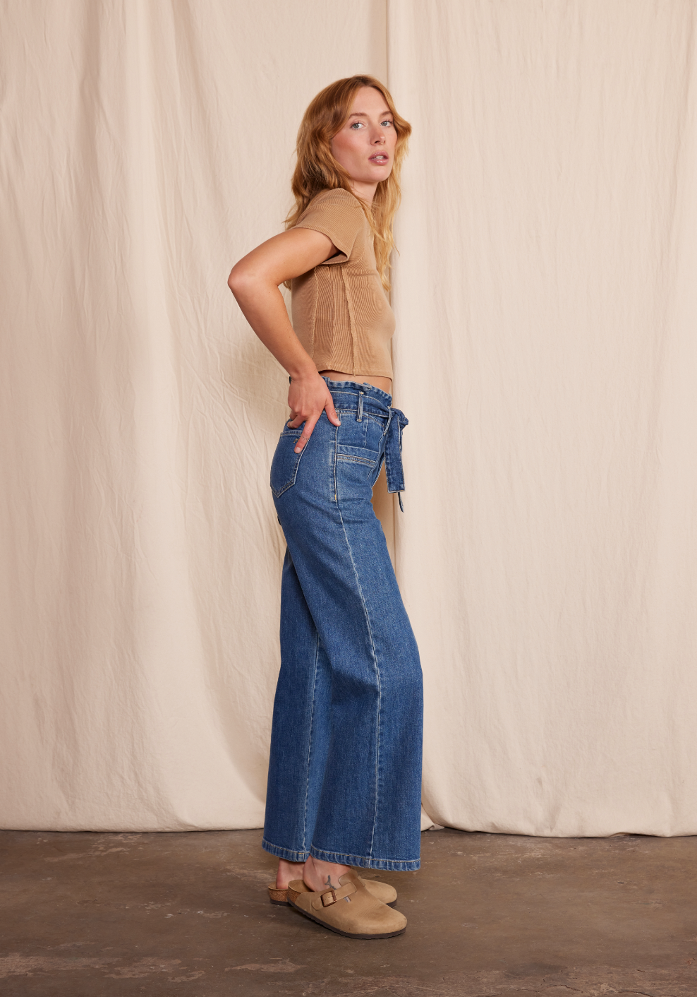 A woman with light brown hair stands in profile against a beige backdrop, wearing Porter Blue Apparels PENNY PAPERBAG WIDE LEG IN LAGUNA WASH—high-waisted organic cotton jeans by Candiani Mills—with a tie belt, tan short-sleeve top, and clogs.