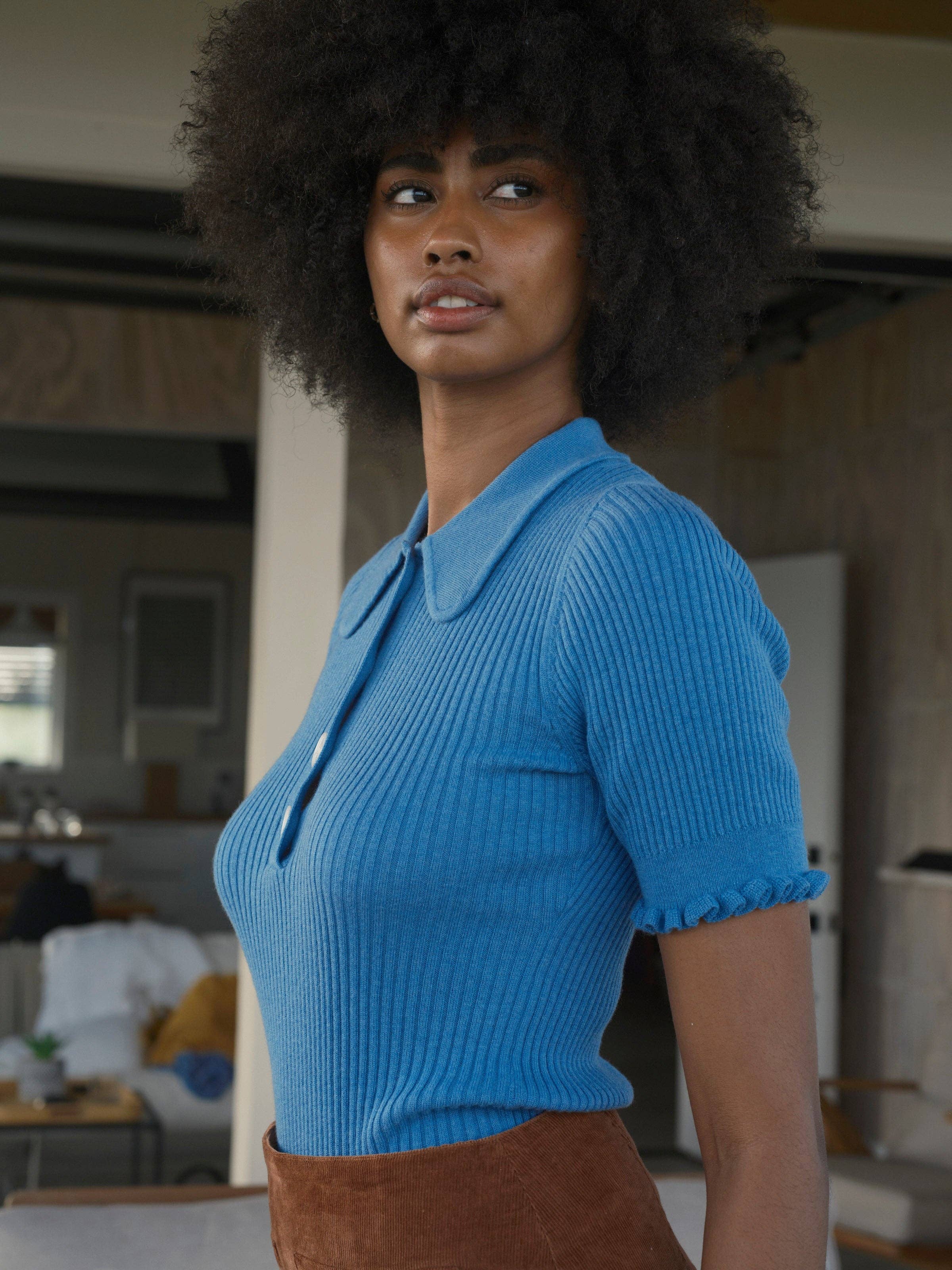 A woman with curly hair wears the vintage-inspired blue Belle Tee by Mollusk with brown pants. Indoors, she looks slightly to the side, her expression neutral.