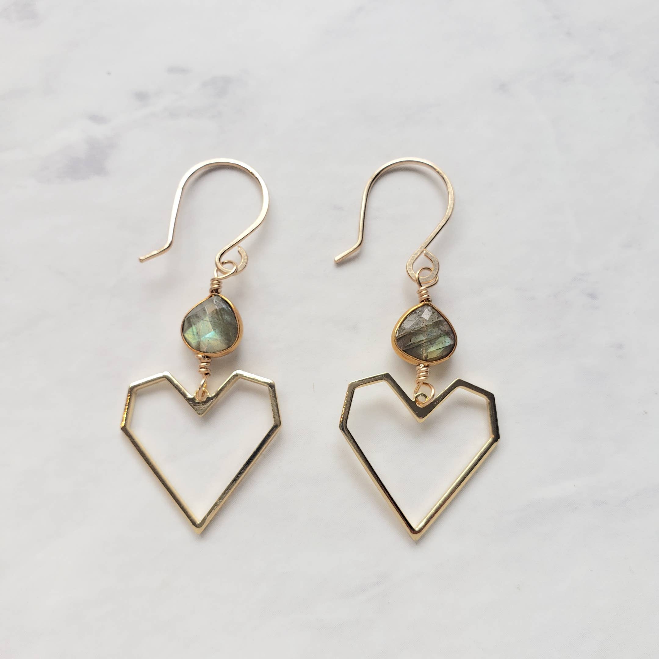 Dreamers of Dreams Geometric Heart Labradorite Earrings feature iridescent labradorite beads above gold-filled geometric hearts, beautifully displayed on a light marble surface.