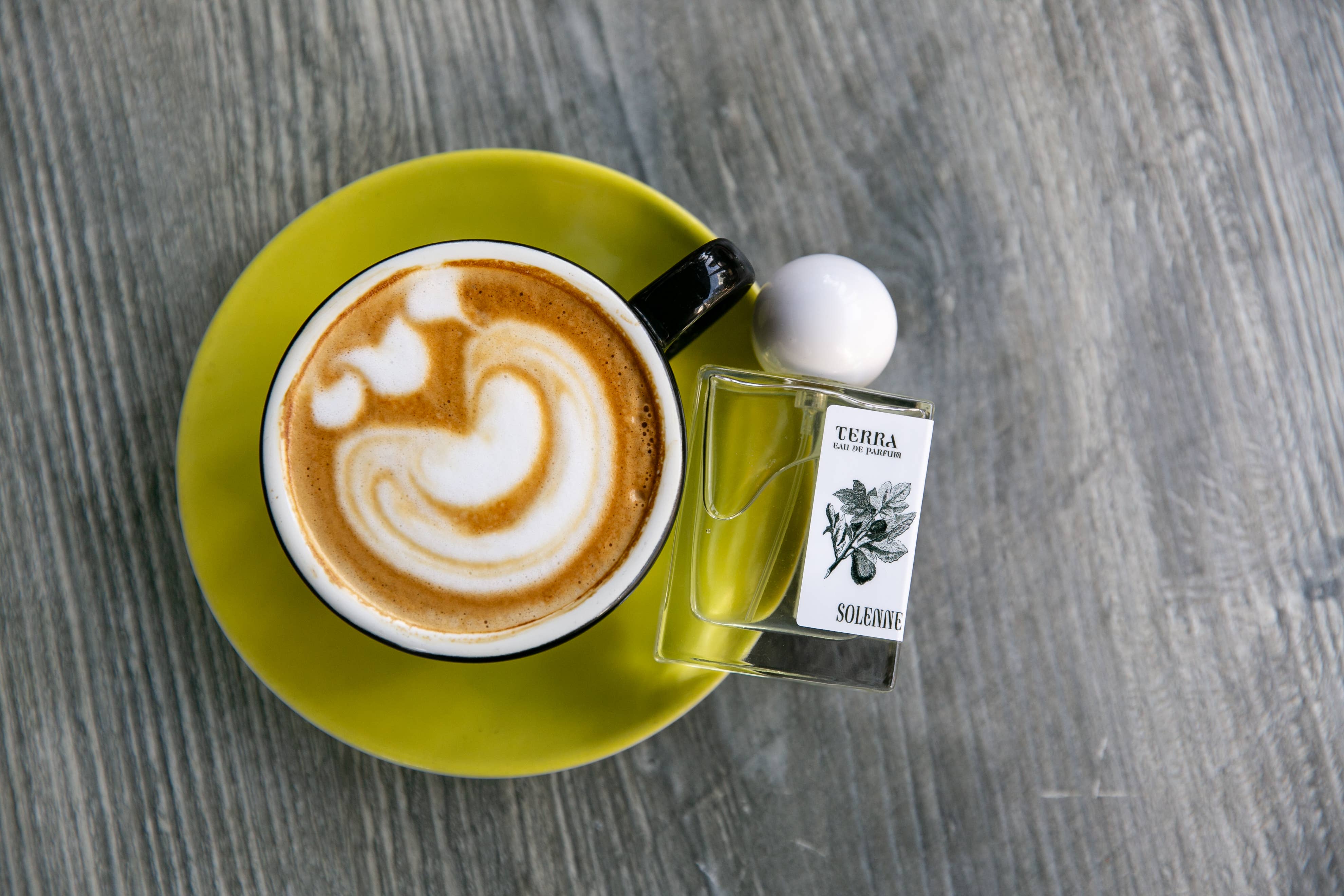 TERRA - Eau De Parfum - Nontoxic Fragrance bottle beside a latte with heart latte art on green saucer on wood table