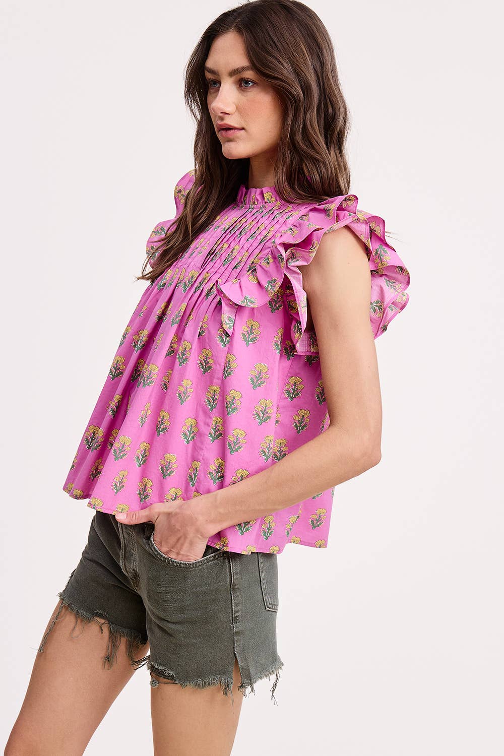 A woman stands sideways in the Ultramarine Celeste Flutter Sleeve Blouse, a lightweight pink floral top, paired with green frayed denim shorts against a plain light background.