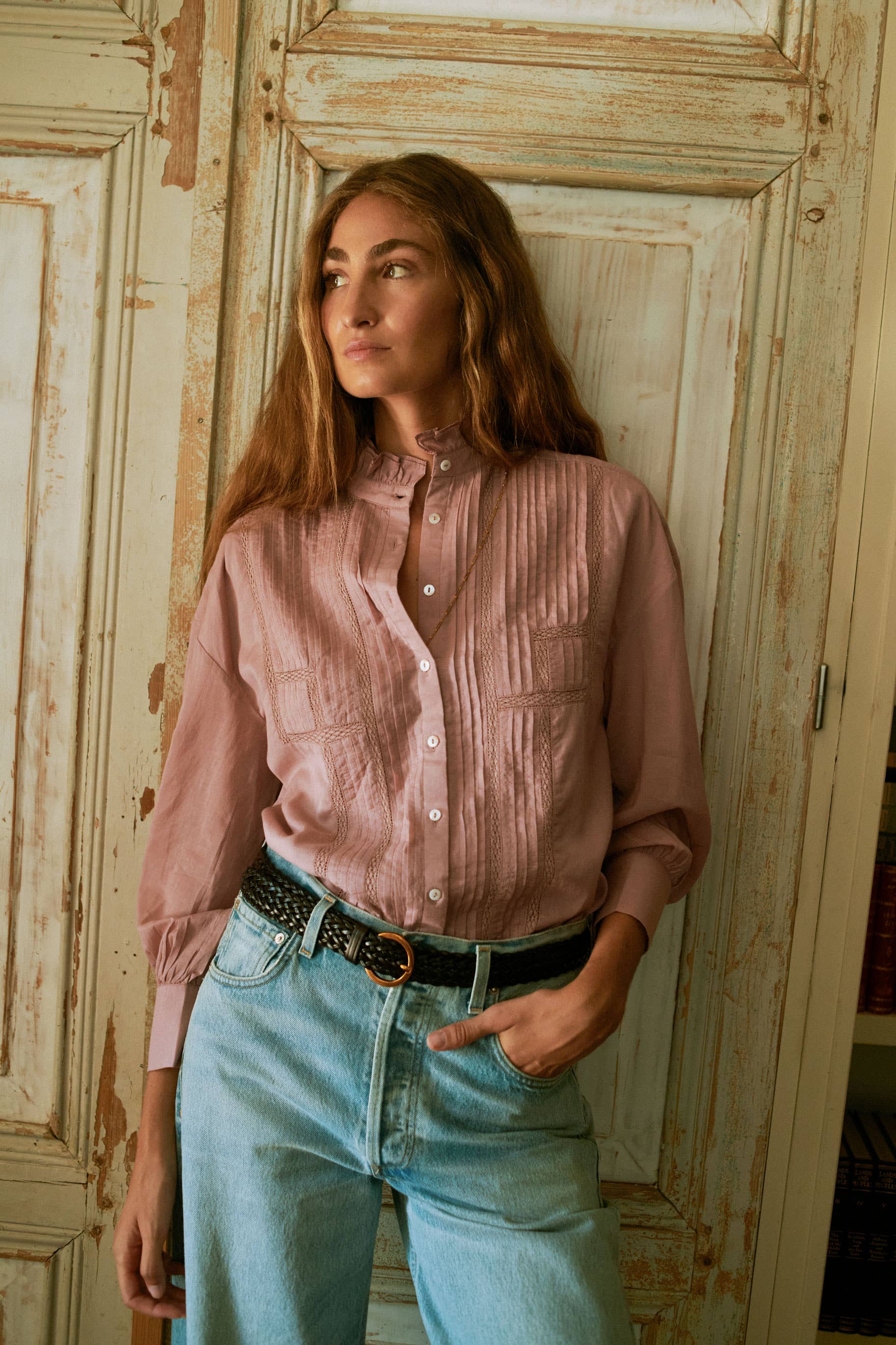 A woman with long brown hair wears the Alexa Blouse Rose by Sonmer Shop USA and light blue jeans, standing against a distressed wooden door with one hand in her pocket—ideal for effortless fall layering.