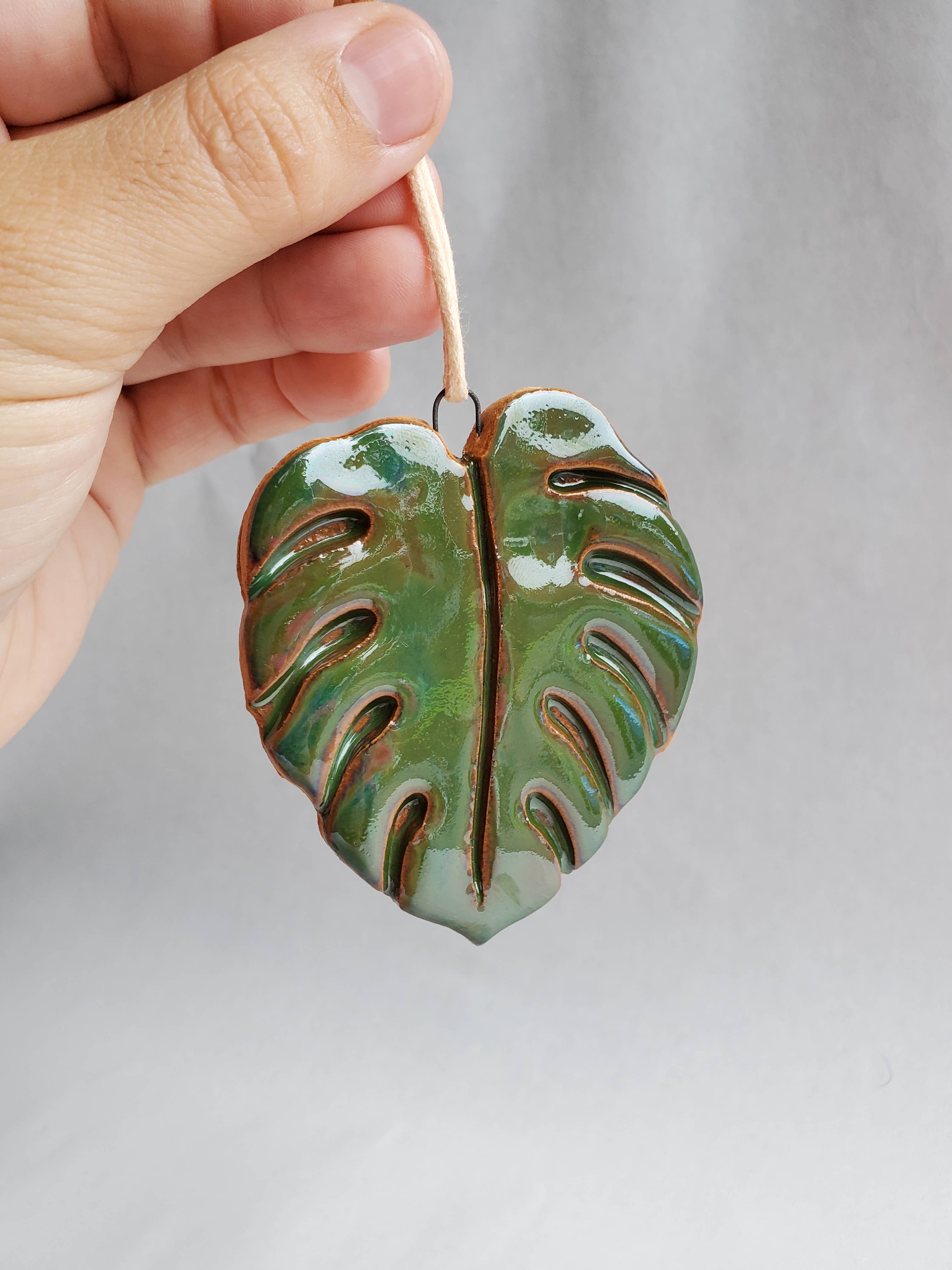 Hand holding glossy green Monstera leaf ornament with a loop string against a plain background
