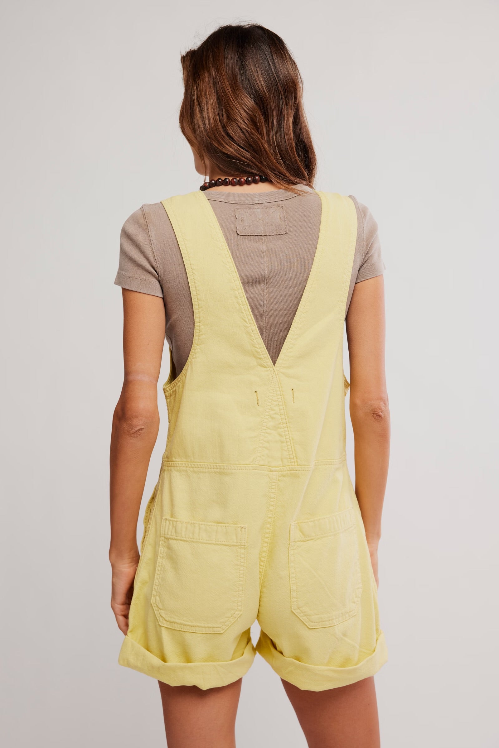 Woman wearing yellow Free People Jude Shortall Overall Short layered over a taupe short sleeve top, back view