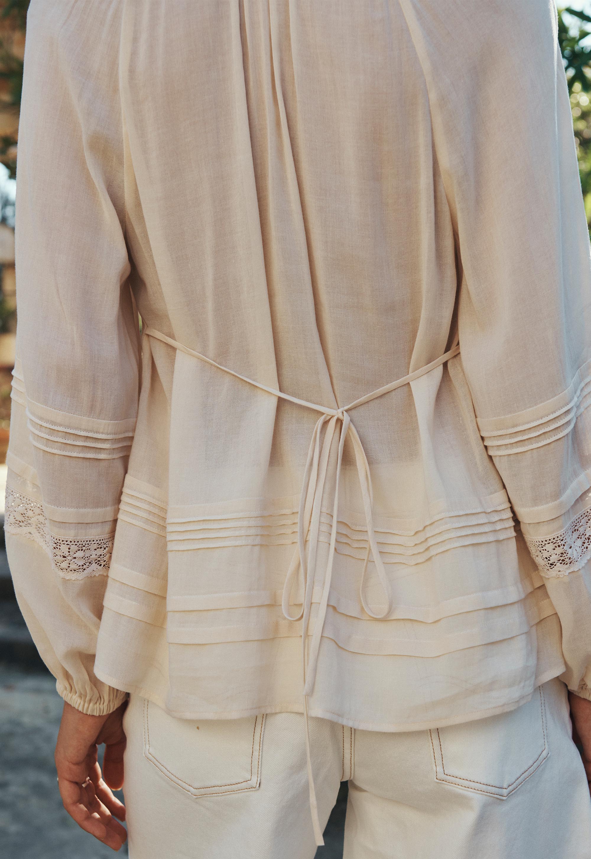 Back view of Hera Lace Blouse- Macadamia with delicate pintuck and lace details, tied at the waist over white pants