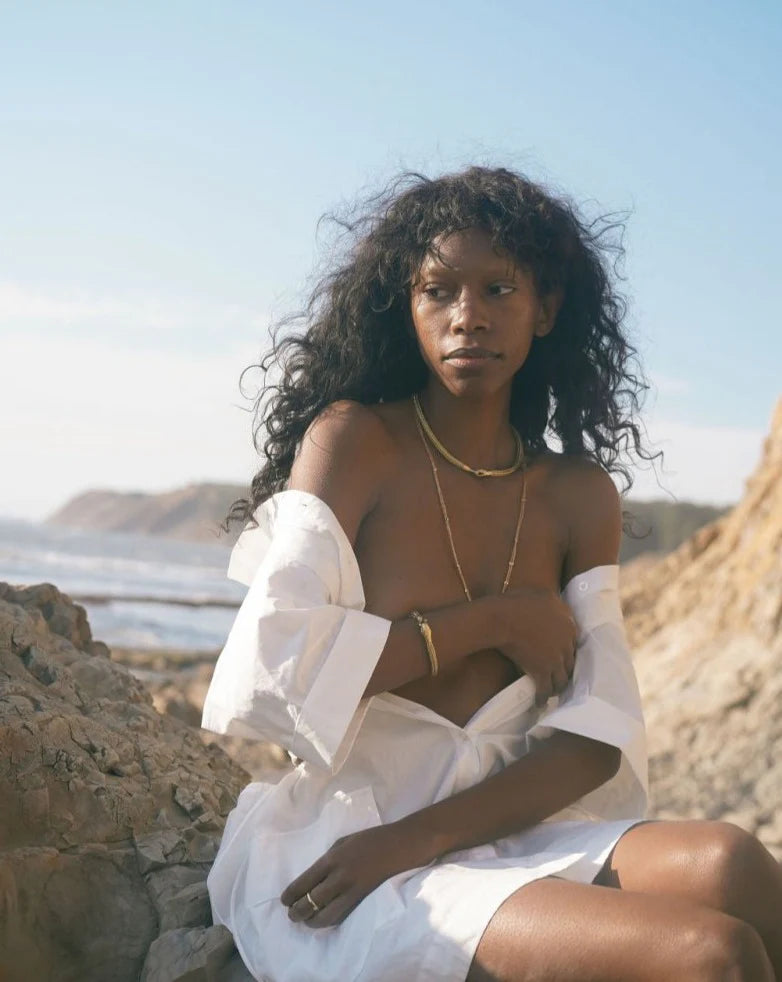 Woman wearing Serpent Choker Necklace with loose white shirt, sitting on rocky beach under soft sunlight