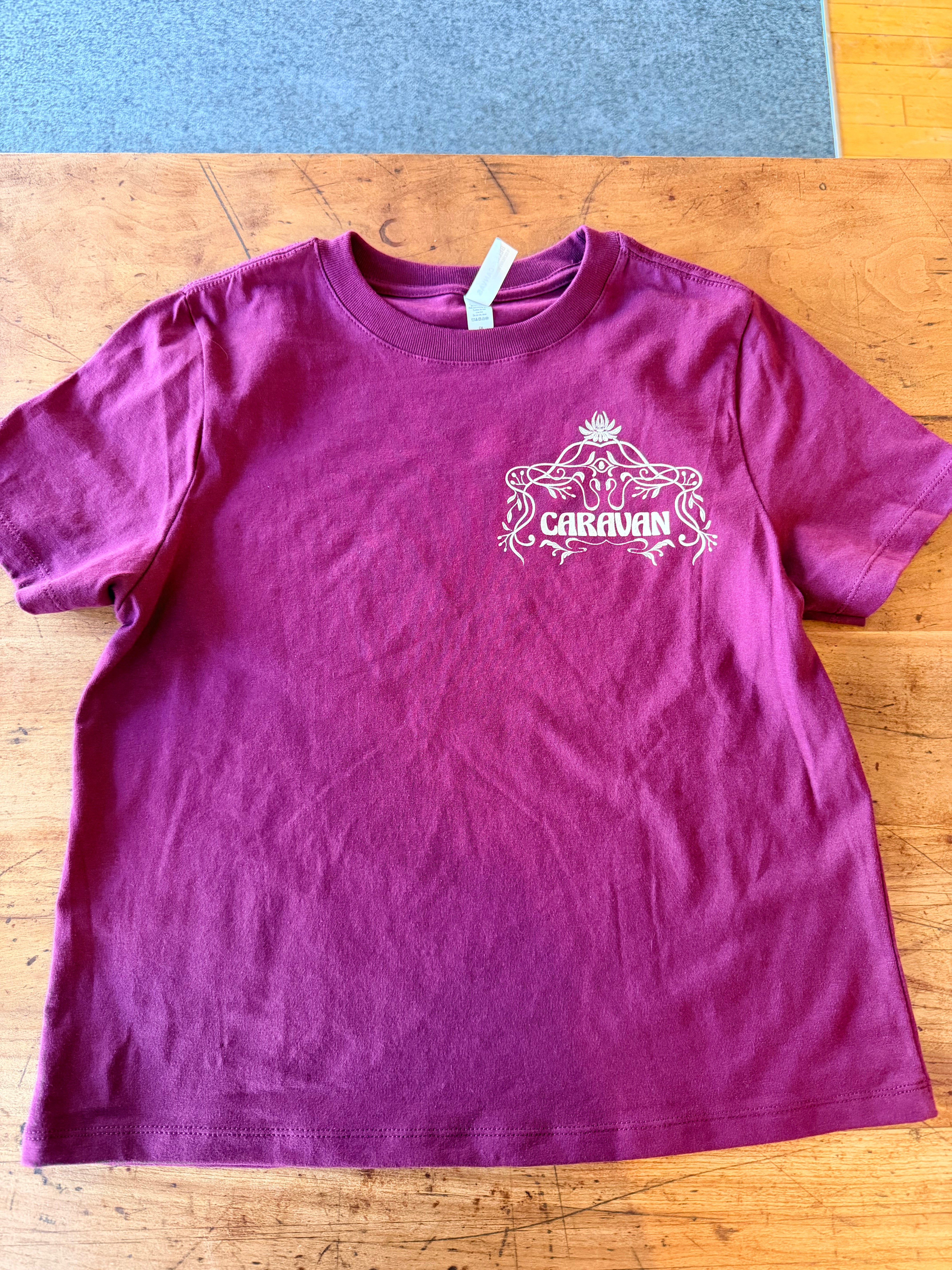 Caravan Legacy Tee in rich burgundy with intricate white graphic design on chest, laid flat on wooden surface.