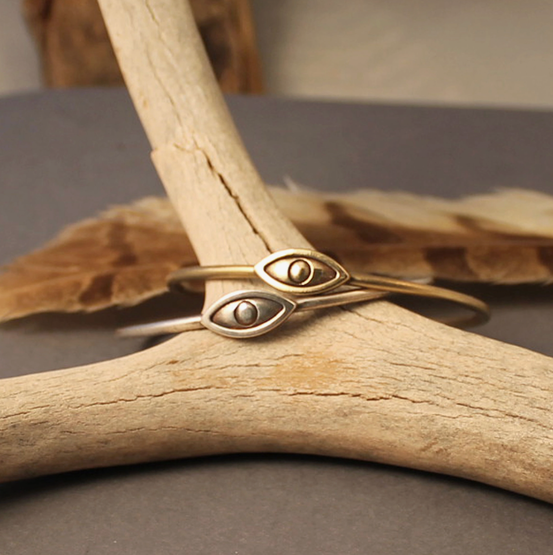 Two Eye Cuff Bracelets in Sterling Silver by Caravan rest on a light-colored antler, with a brown feather in the background on a dark surface.