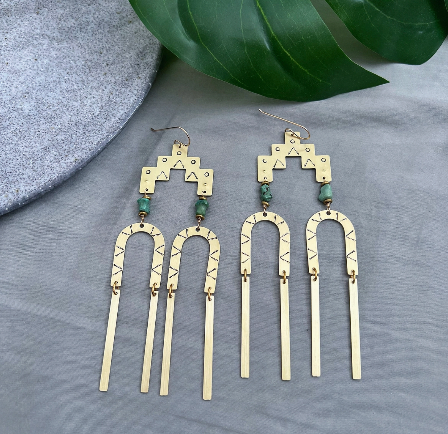Brass Dangle Earrings by Lunasol—featuring geometric shapes, etched details, and green beads—are striking handmade earrings displayed on gray near a stone slab and green leaf, perfect for unique jewelry lovers.