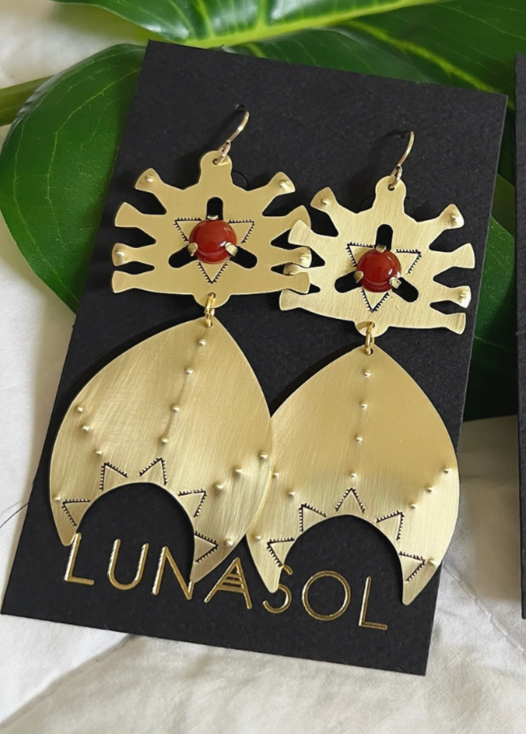 The Astra Earrings in Carnelian or Mother of Pearl by Lunasol for Caravan feature intricate brass cut-outs, red beads, and engraved patterns with gold-filled earwires, shown on a LUNASOL card with green leaves in the background.