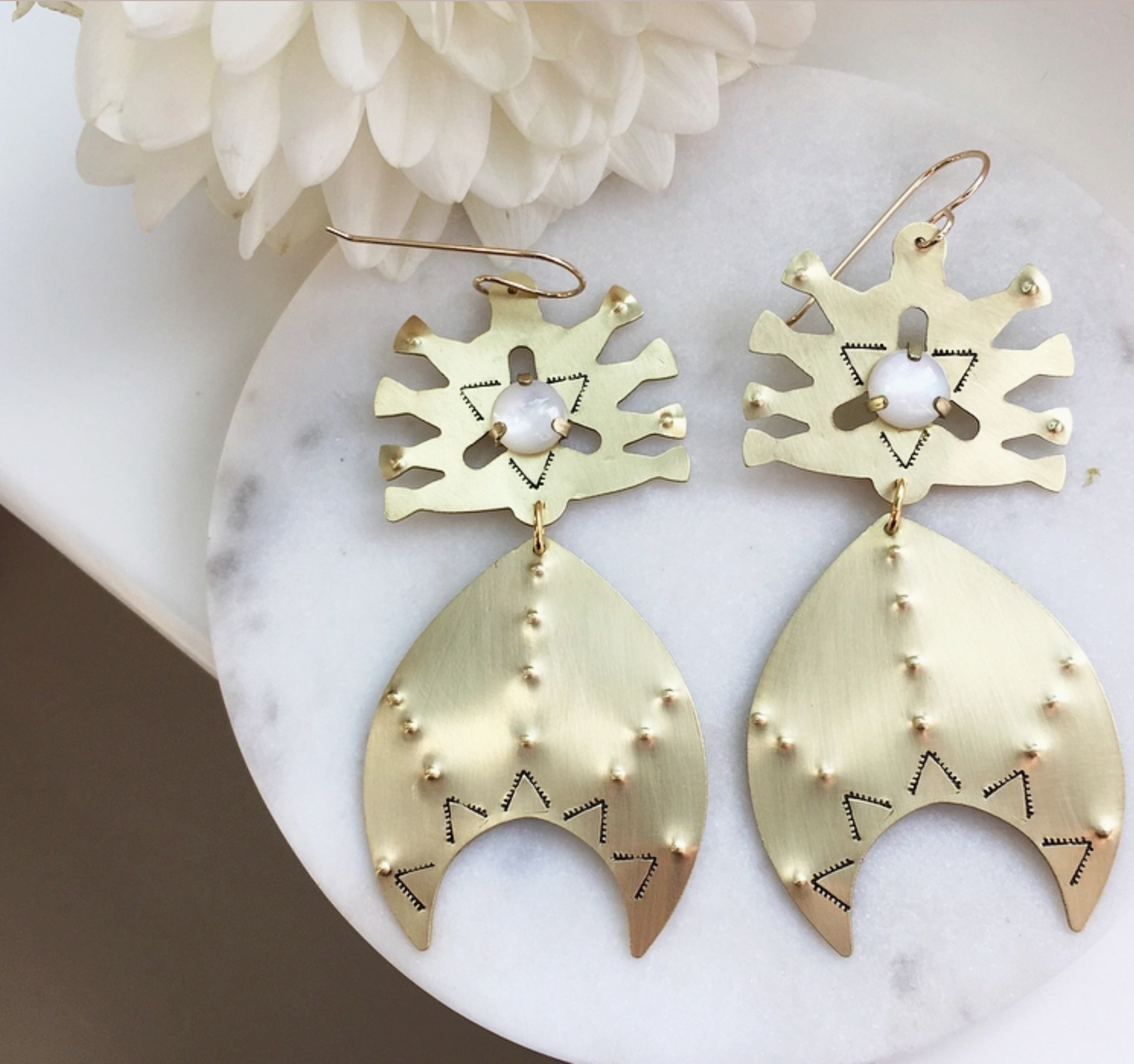 The Astra Earrings in Carneian or Mother of Pearl by Lunasol for Caravan feature geometric gold dangles with abstract shapes and pearl accents, shown on a marble surface beside a white flower.