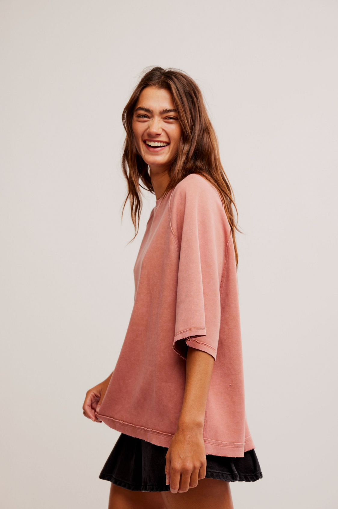 A young woman with long brown hair smiles while standing sideways. She wears the Joah Tee in Cedarwood by Free People with elbow-length sleeves and a short black skirt, set against a plain, light-colored background.