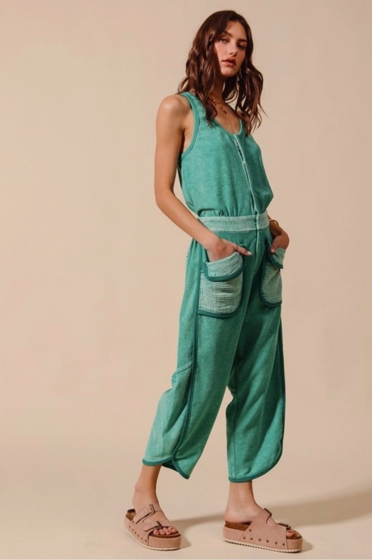 A woman poses against a beige background wearing Caravan’s Mineral Washed Button Up Scoop Neck Terry Jumpsuit in teal-green, featuring large light-green front pockets, paired with beige platform sandals. She has wavy brown hair and hands in her pockets.