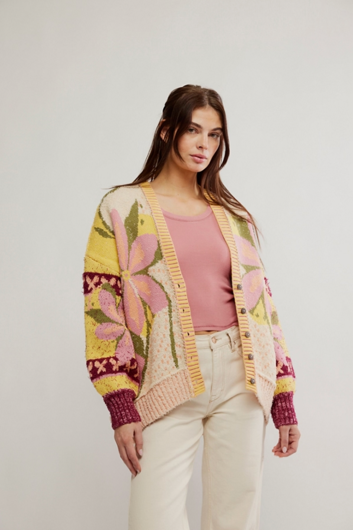 A woman with long brown hair wears the Bali Tropical Garden Sweater by Free People, an oversized cotton-blend cardigan with large pink and yellow flowers, paired with a pink top and cream pants against a plain light background.