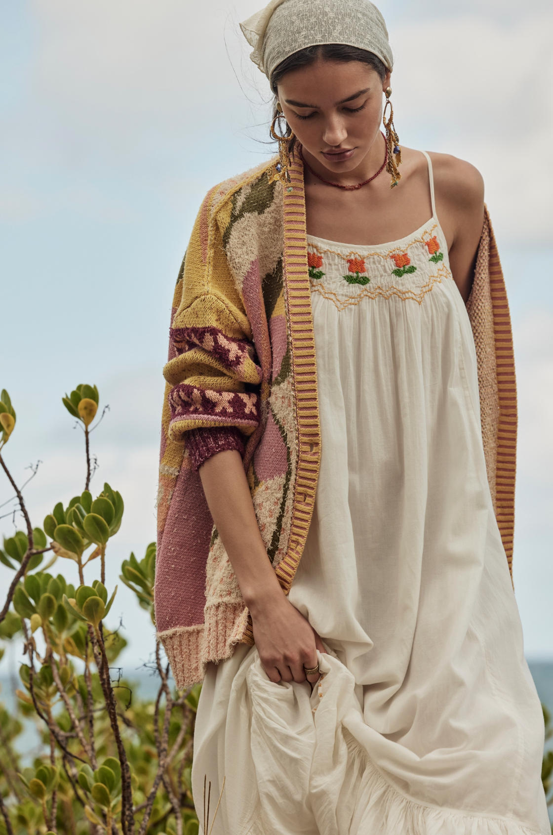 A woman stands outdoors in the Bali Tropical Garden Sweater by Free People, paired with a white embroidered dress, headscarf, and earrings. Greenery and a cloudy sky complete the background.